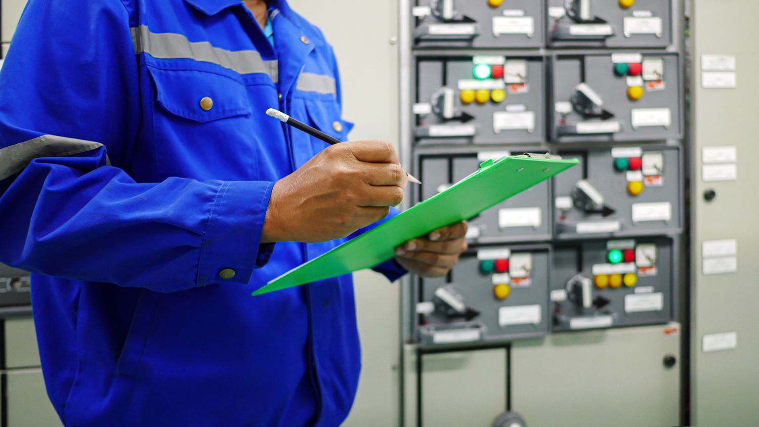Technician in blue coveralls holding a green clipboard in front of electrical control panels Technician in blue coveralls holding a green clipboard in front of electrical control panels