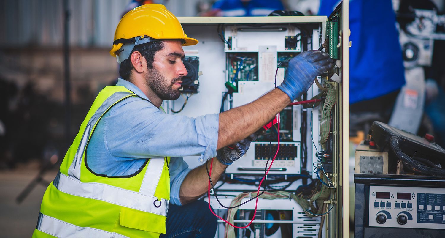 Technician in yellow hard hat and vest inspecting wiring inside an electrical control panel