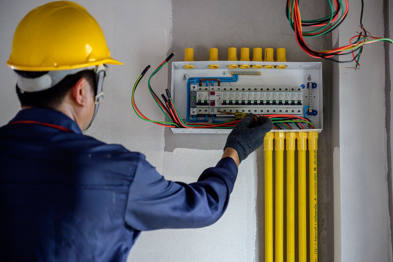 Electrician in a yellow hard hat inspecting an open circuit breaker panel with exposed wires. Electrician in a yellow hard hat inspecting an open circuit breaker panel with exposed wires.