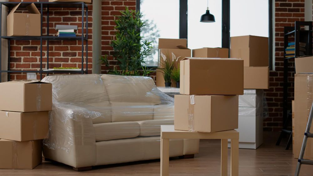 Moving boxes stacked around a wrapped sofa in a room with exposed brick and a window.