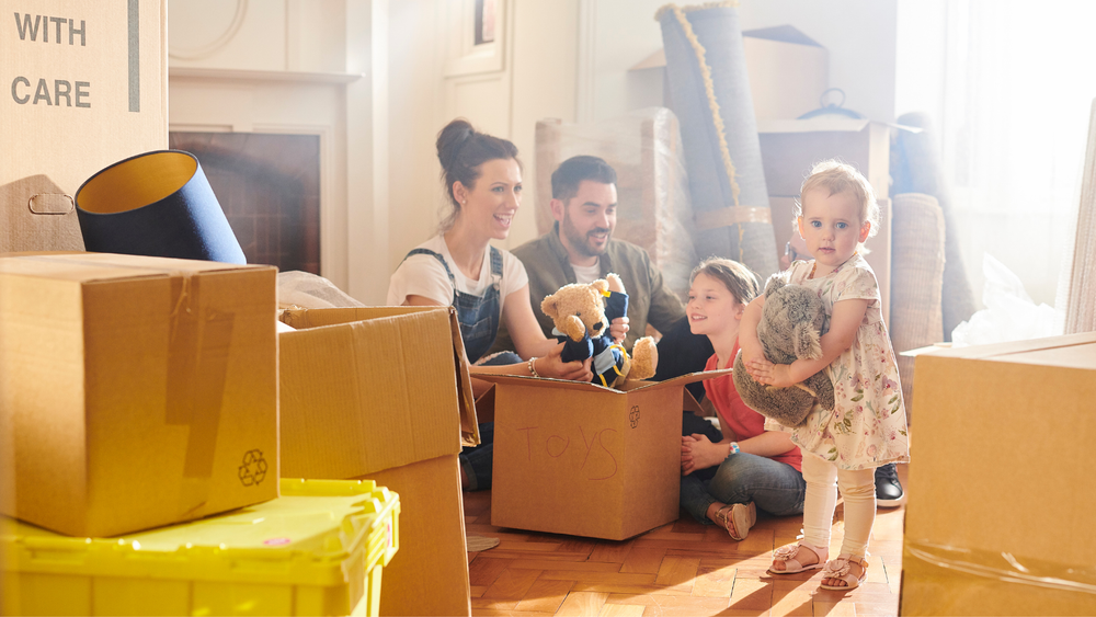 Family unpacking boxes in a room, with a little girl holding a stuffed animal.