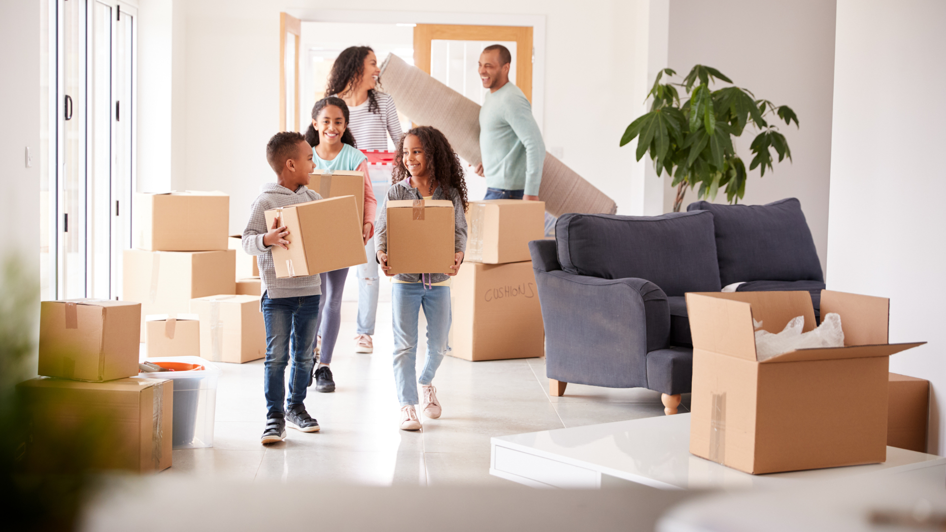 Family carrying boxes and a rolled rug into a bright, empty living room with a sofa and plant.