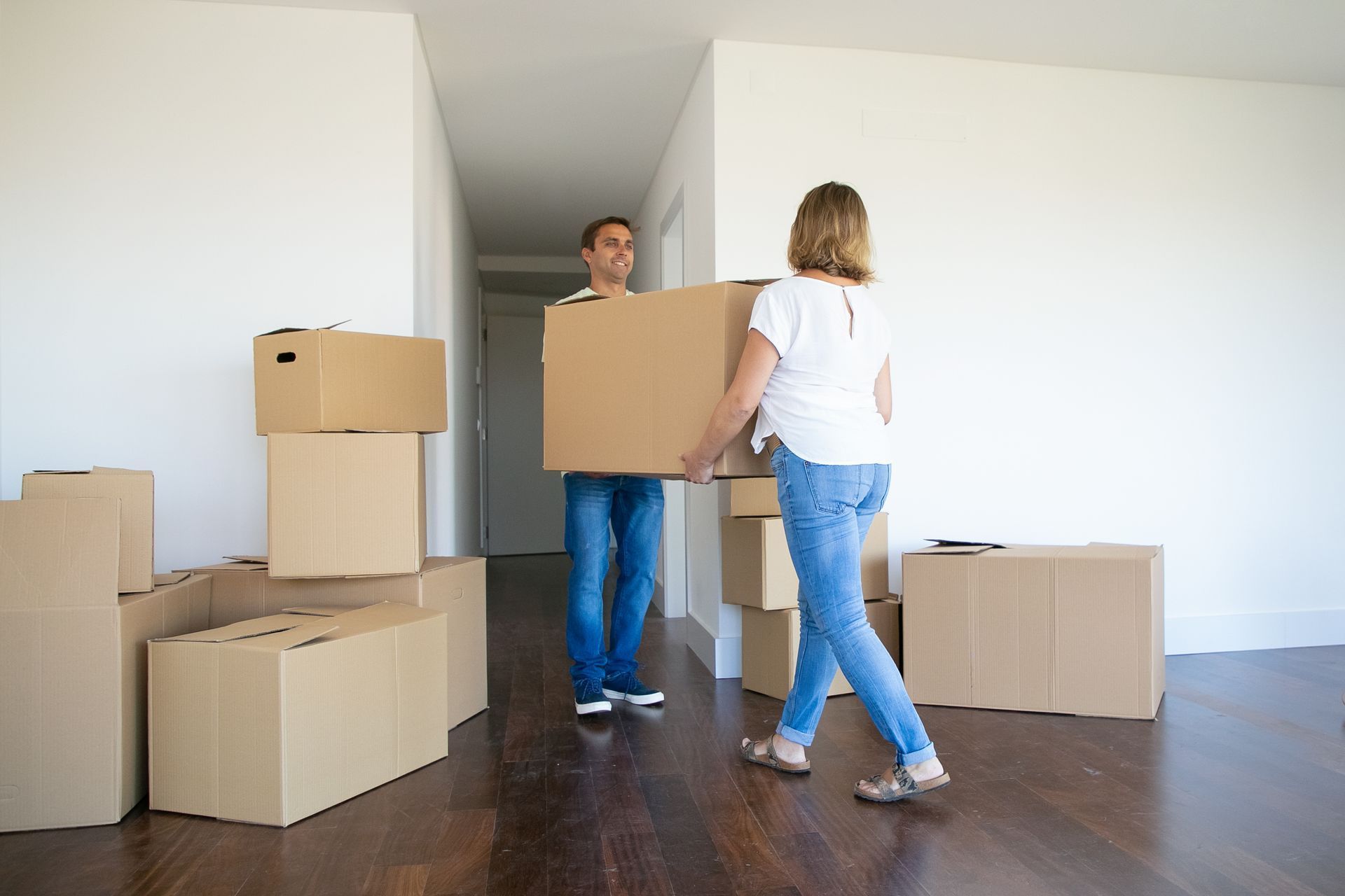 Couple carrying a large cardboard box in a new, empty apartment with other boxes around.