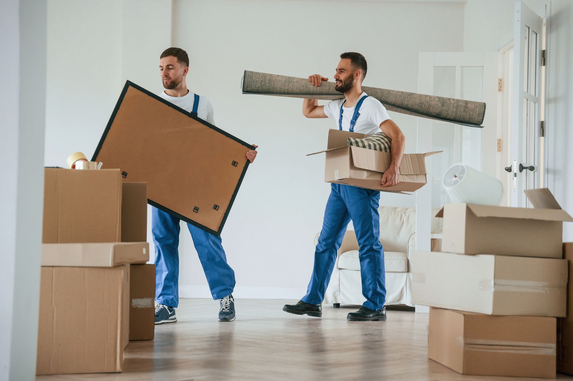 Two movers in blue coveralls carrying packed boxes, a large framed item, and a rug in a bright room.