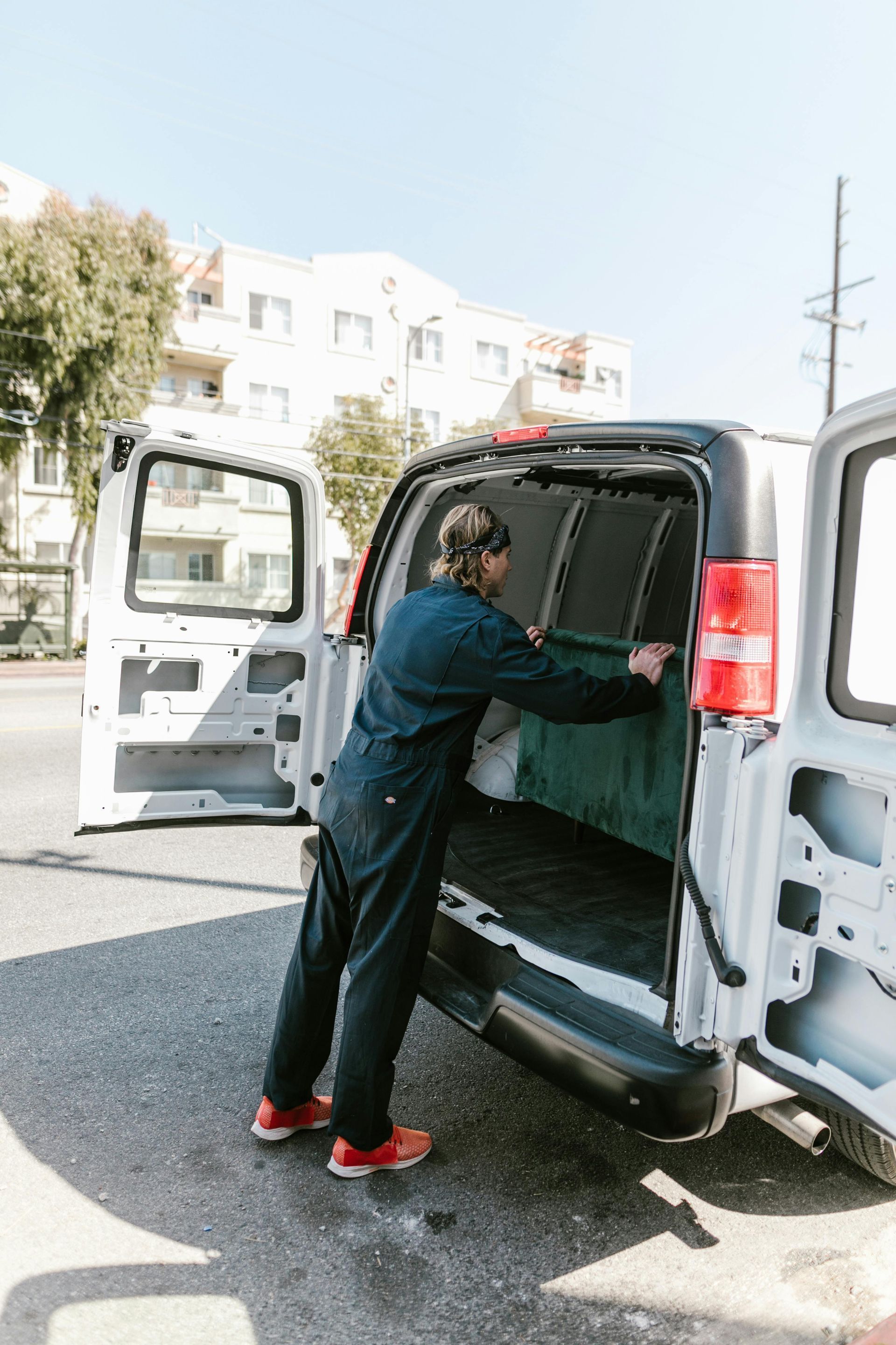 Person in navy jumpsuit loading the back of a white van with the doors open on a city street.