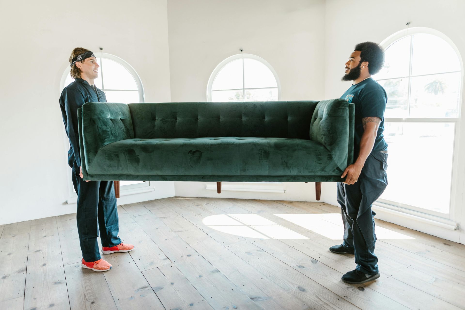 Two people carrying a green velvet sofa in a bright room with arched windows.