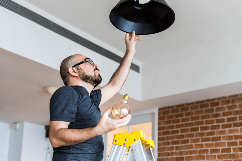 Man on a ladder replacing a light bulb in a black pendant lamp, in a room with exposed brick and white walls.