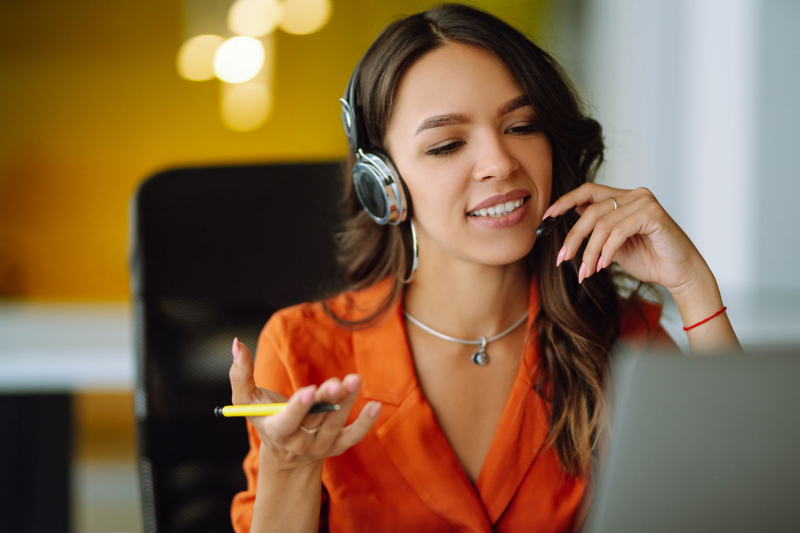 Woman in orange blazer wearing headphones, gesturing, and looking at laptop screen.