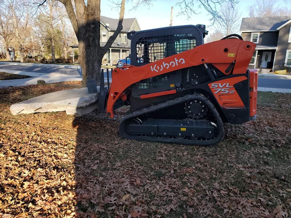 A kubota skid steer is parked in a yard next to a tree.