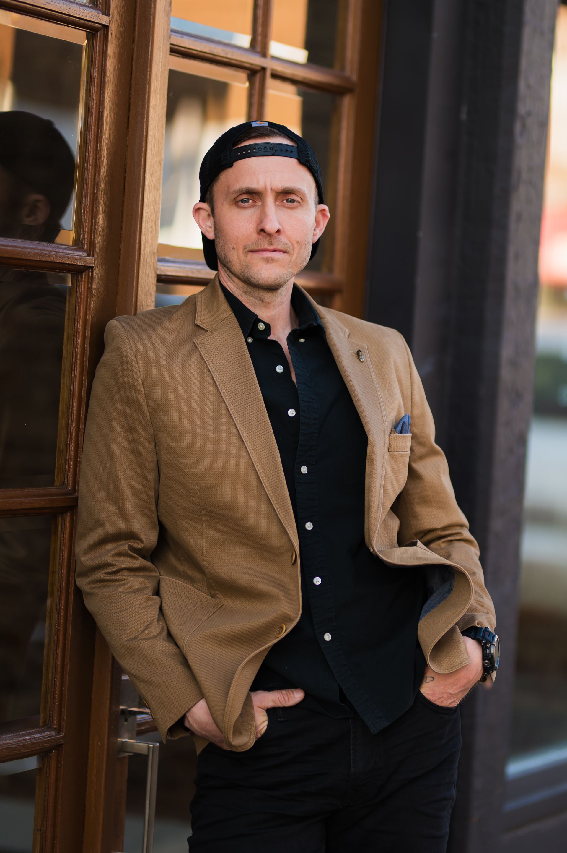 Man in blazer and cap, leaning against a wooden door, hands in pockets, looking at the camera.