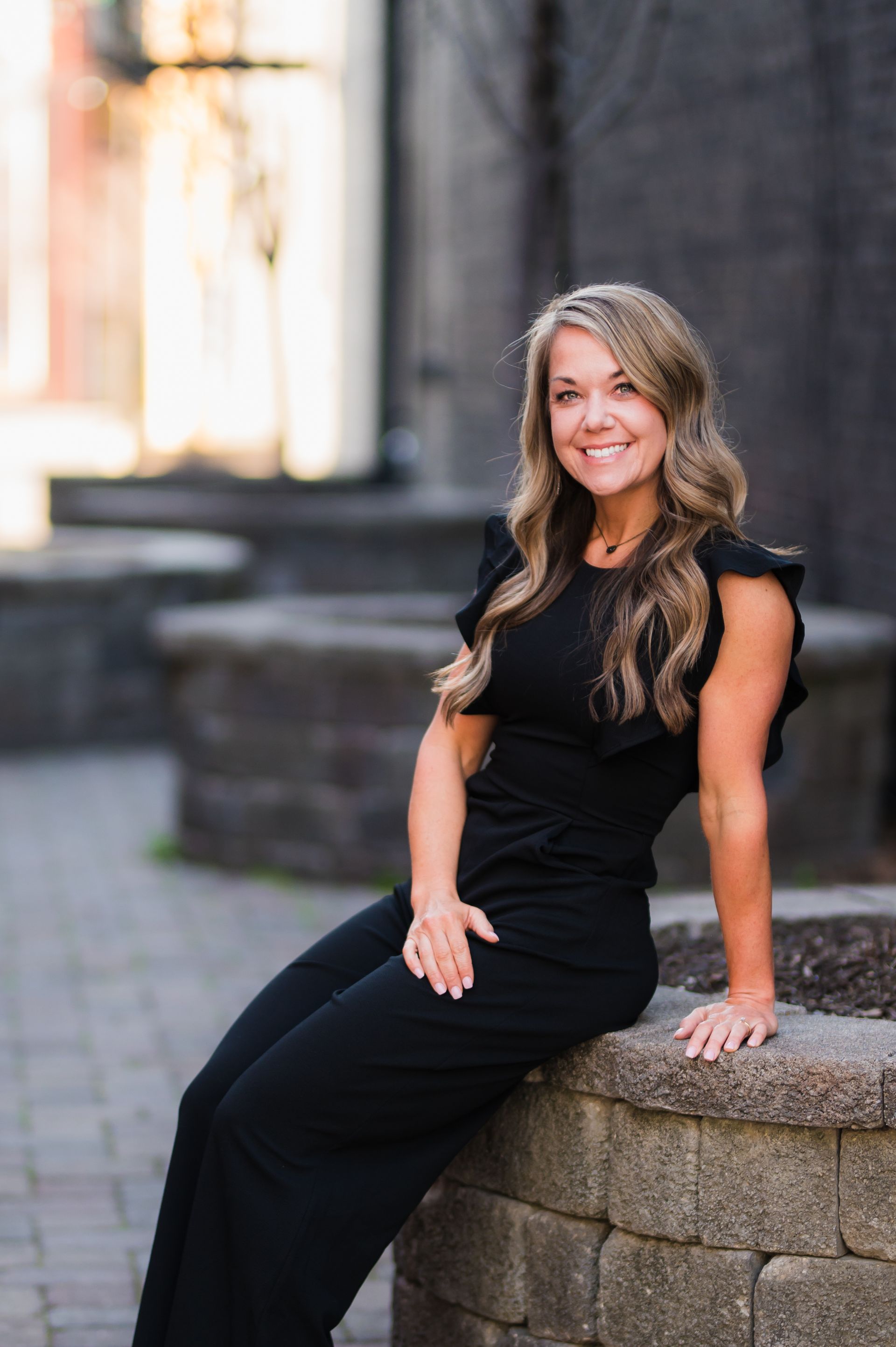 Woman in black jumpsuit, smiling, sitting on a stone wall outdoors.
