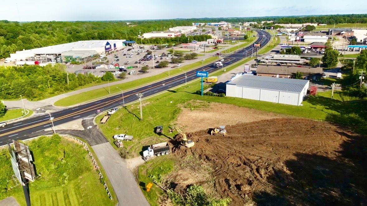 Aerial view of a road with businesses; dirt field in foreground, construction vehicles working.