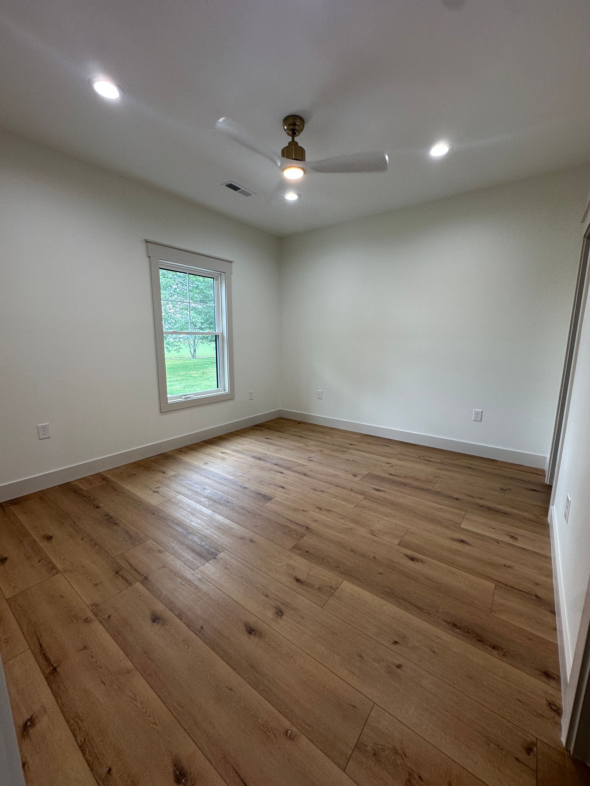 Empty bedroom with wood floors, white walls, window, ceiling fan, and recessed lighting.