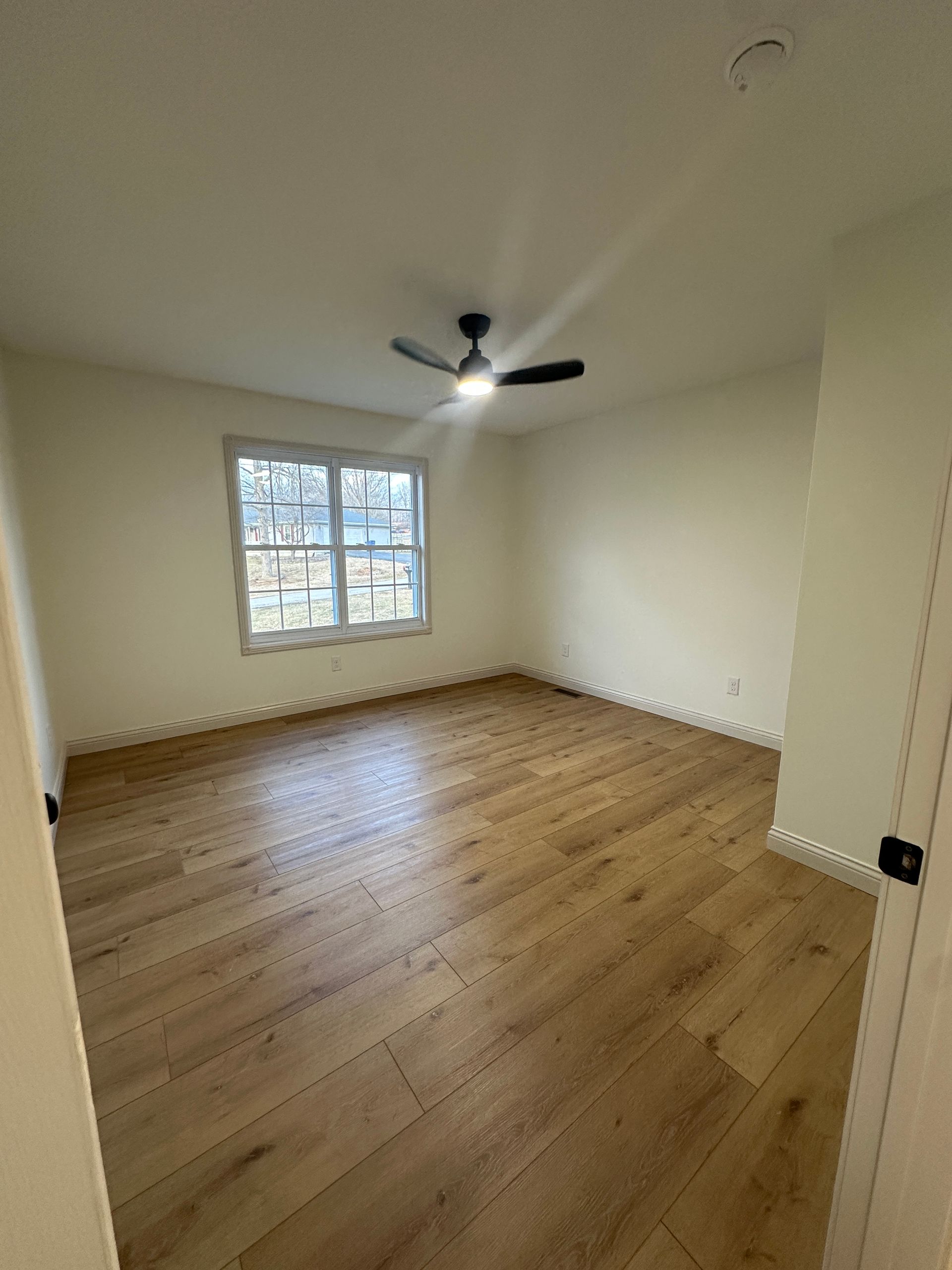 Empty bedroom with wooden floor, window, ceiling fan, and light walls.