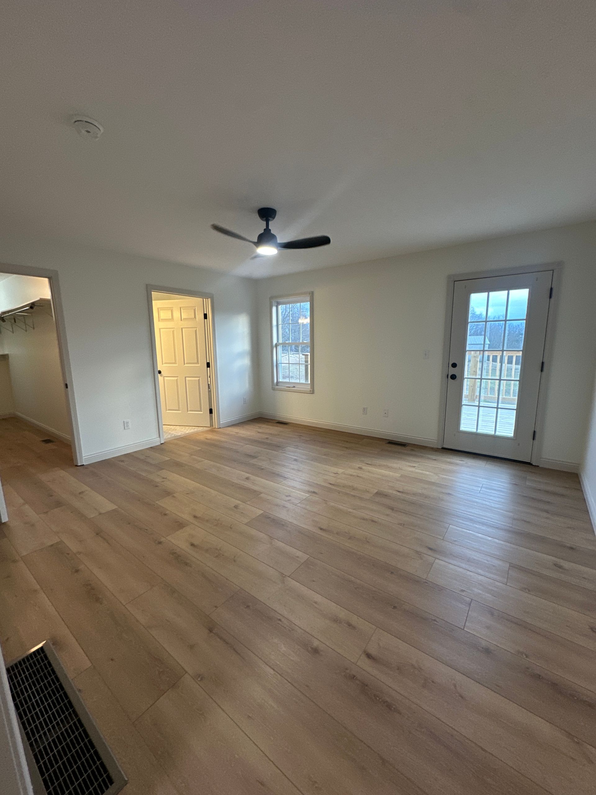 Empty living room with wood flooring, white walls, and a black ceiling fan. A door and windows provide light.