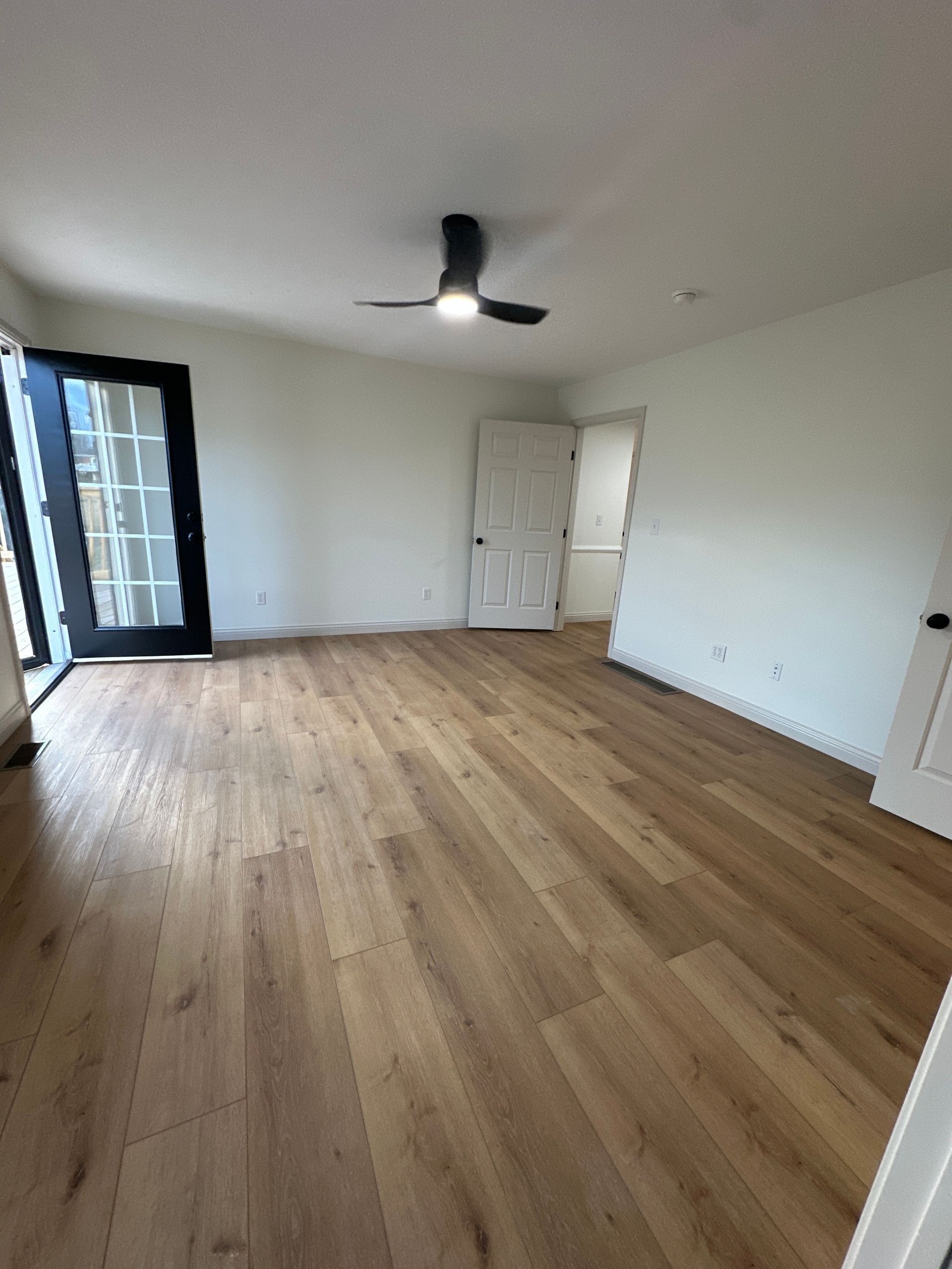 Bright room with wood floors, a black ceiling fan, and french doors. A white door leads to another room.