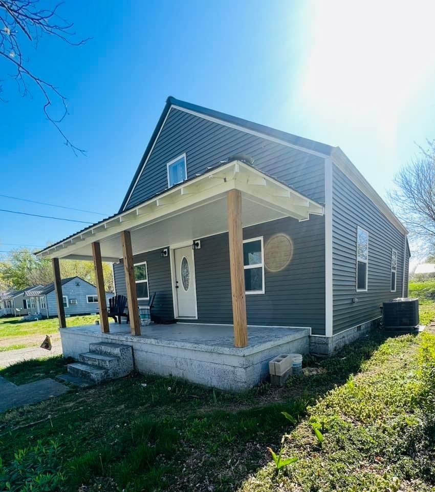 Gray two-story house with a porch, brown columns, and a concrete foundation under a bright blue sky.