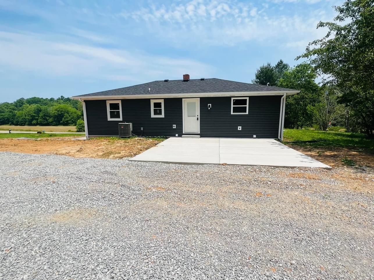 Small, dark-sided house with white trim, door, and windows, set on a concrete slab and gravel driveway, under a blue sky.
