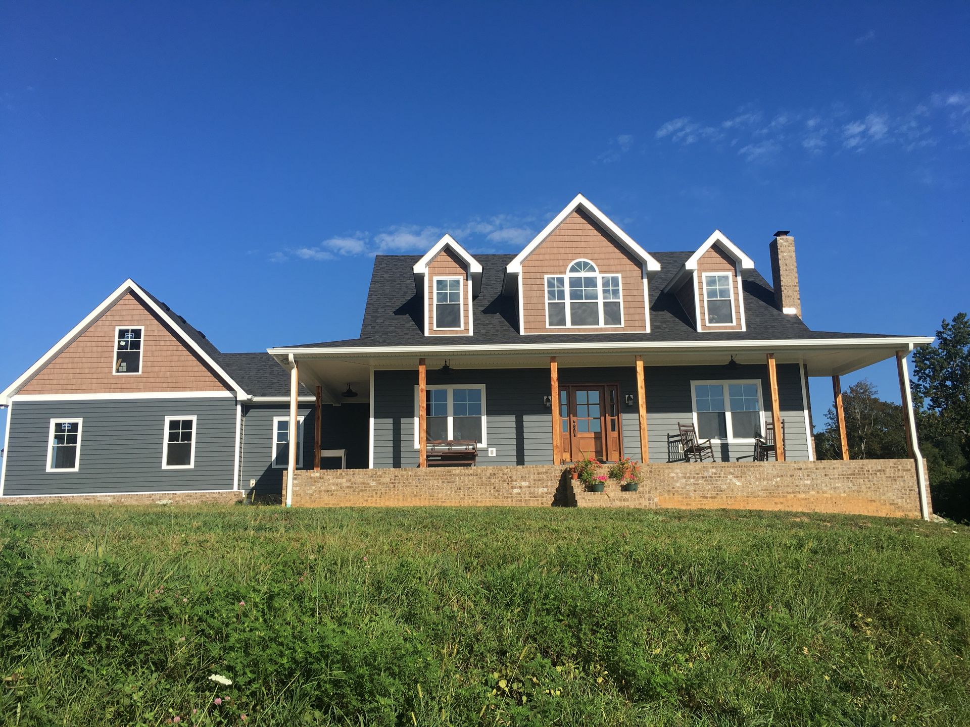 Gray house with brown accents and porch, set on a grassy hill under a bright blue sky.