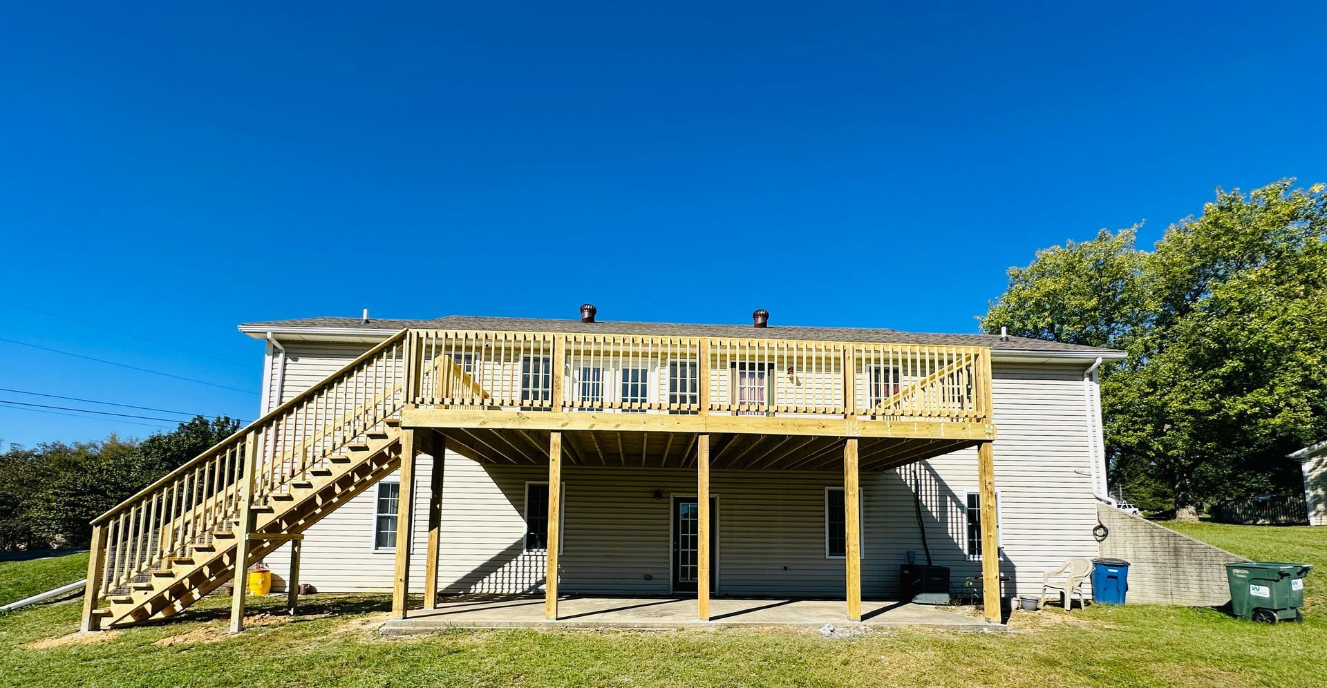 Two-story house with a large wooden deck, stairs, and a concrete patio. Sunny day, blue sky.