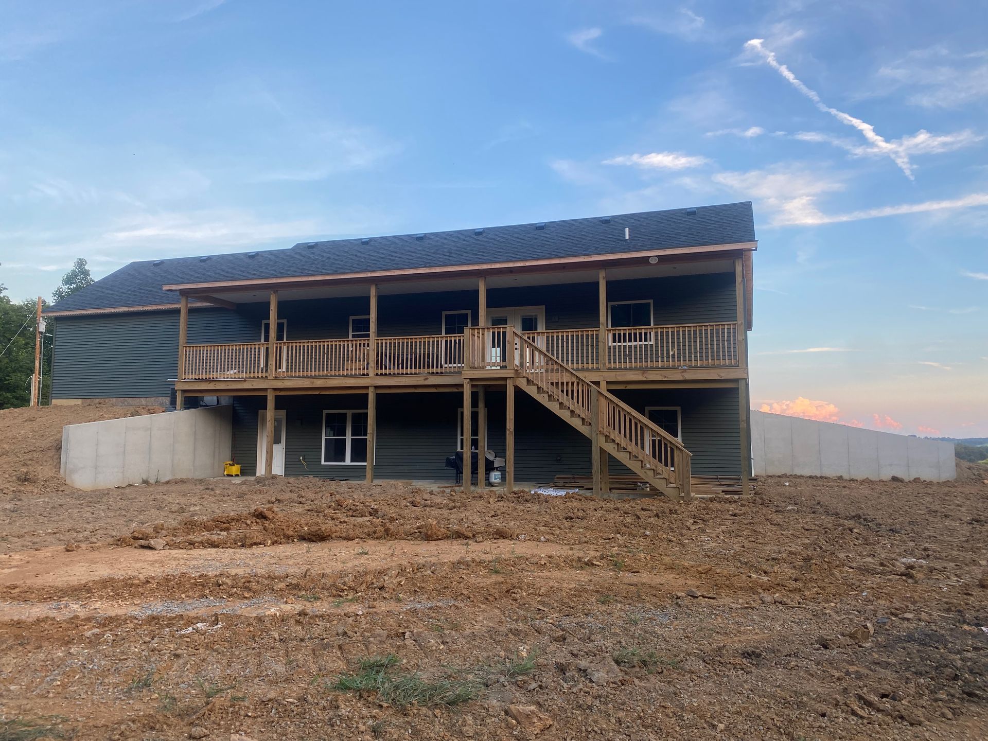 Two-story house under construction with a large wooden deck, set on a hillside with exposed dirt.