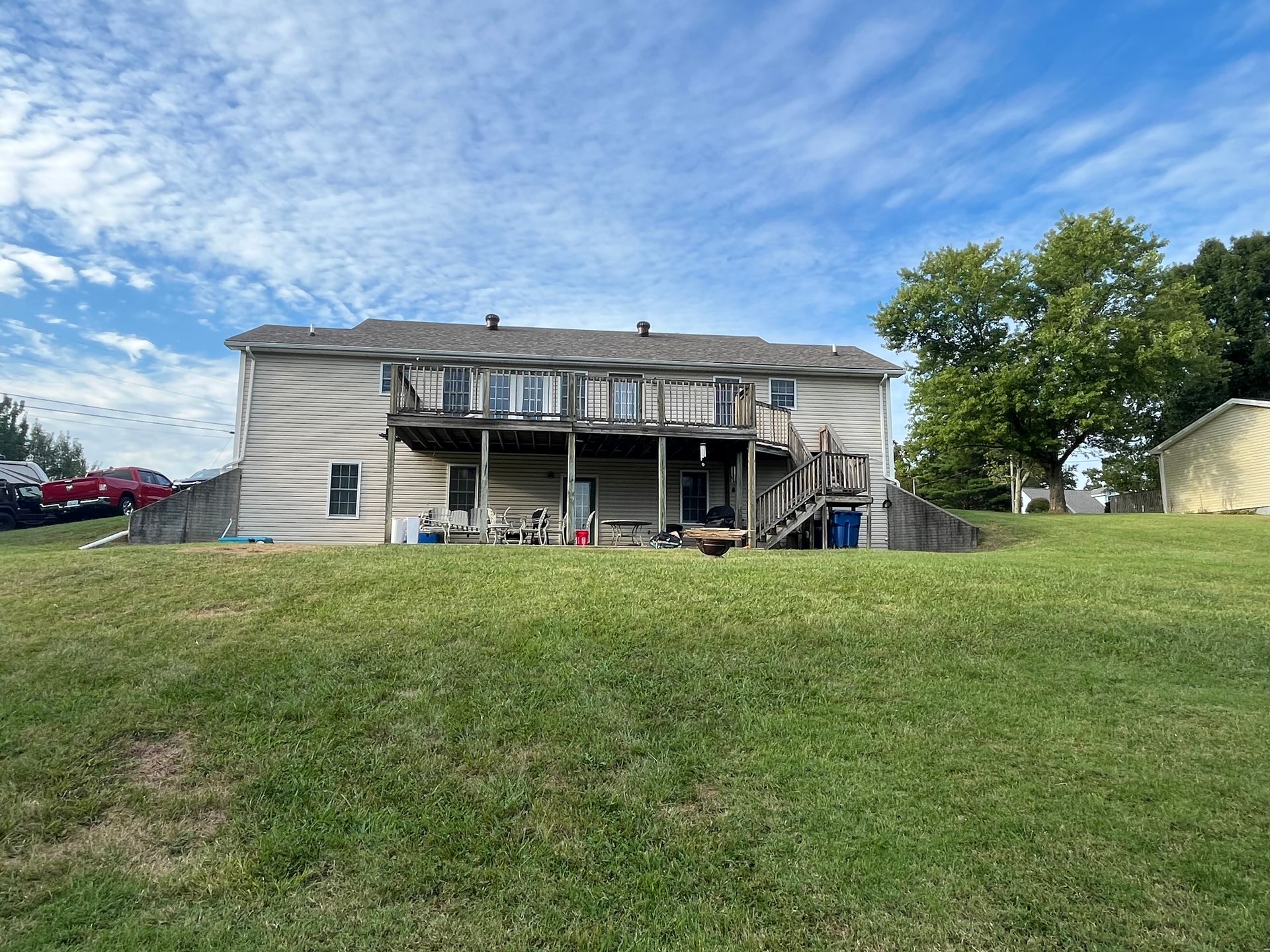 Two-story house with a weathered deck, set on a grassy hill under a blue sky.