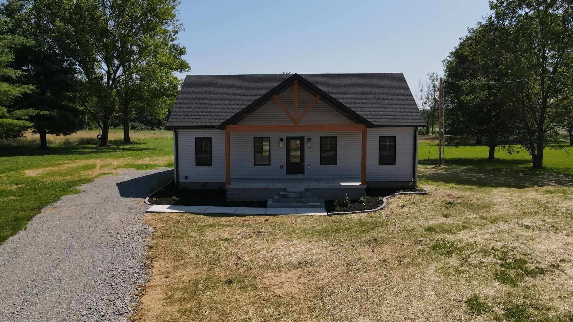 A small, gray house with a wooden porch and a gravel driveway, set in a grassy field.