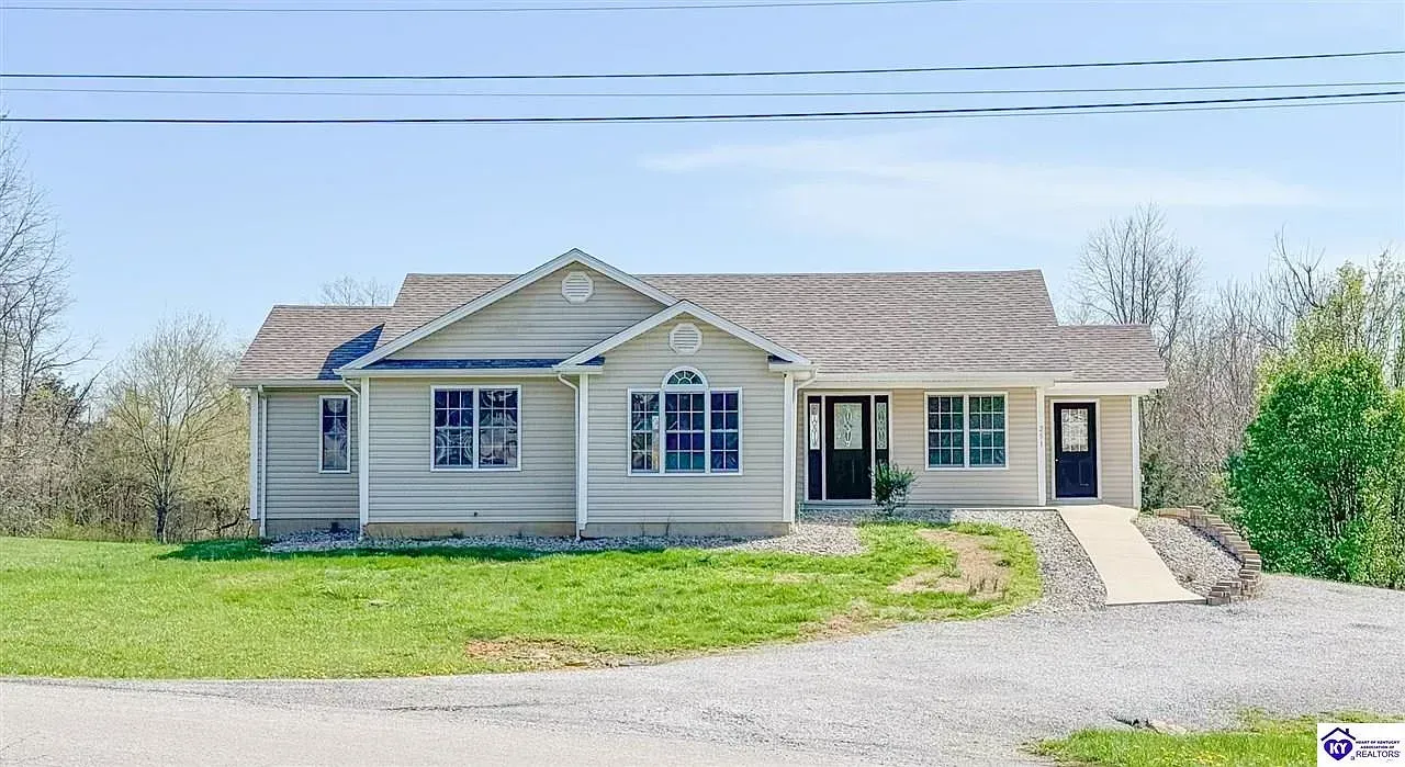 Small, light-colored house with a ramp and gravel driveway, set on a grassy hill under a blue sky.