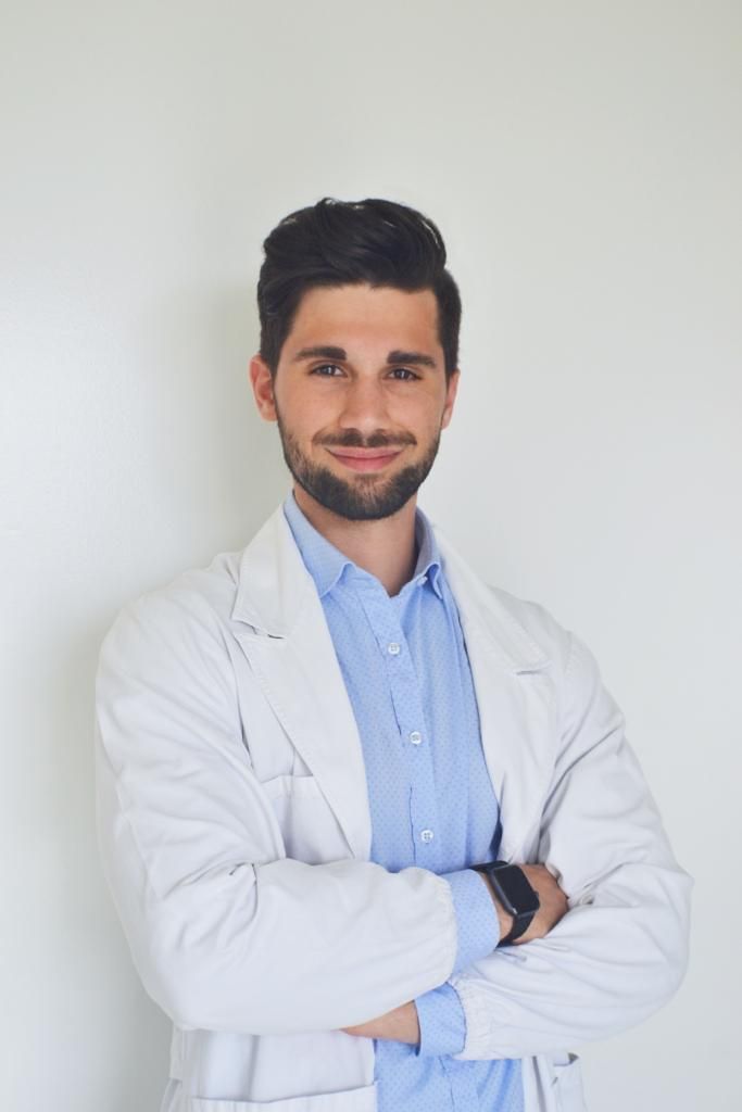 Un uomo in camice bianco e camicia blu è in piedi con le braccia incrociate.