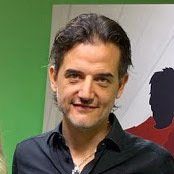 Un uomo con una camicia nera sorride davanti a un muro verde.