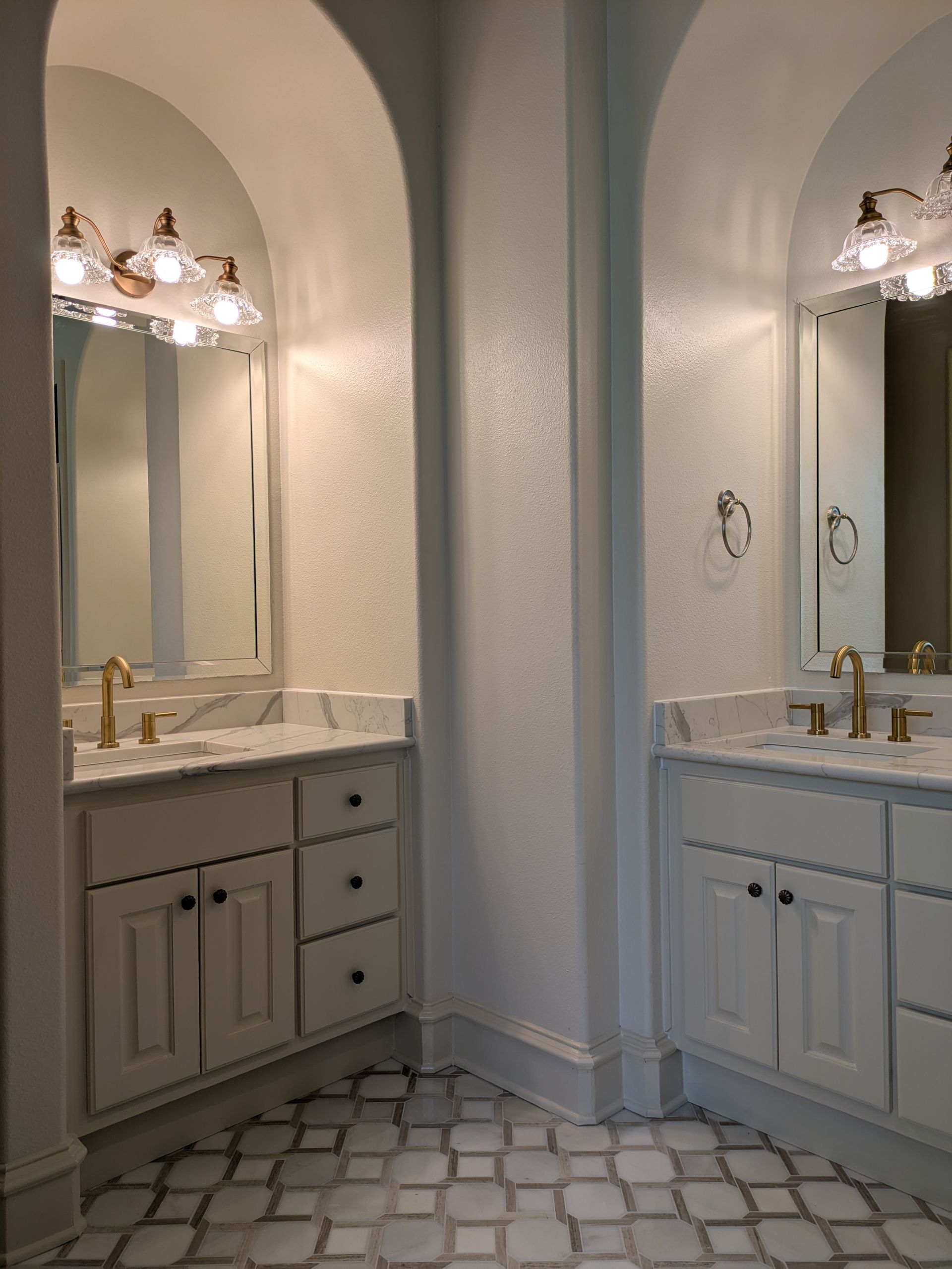 Two white bathroom vanities under arched alcoves with marble countertops and gold fixtures.