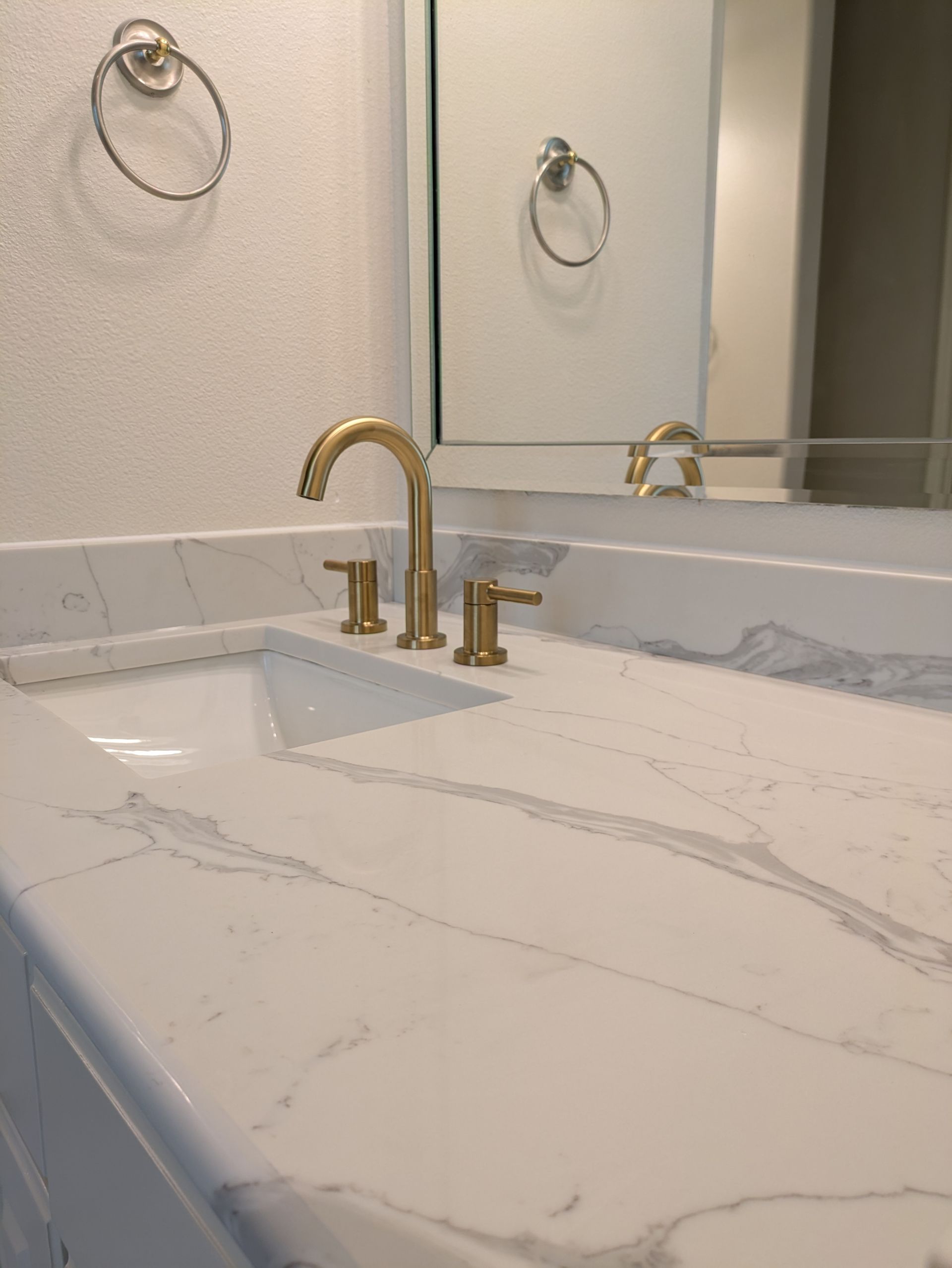 Gold faucet on white countertop with a square sink and mirror reflection.