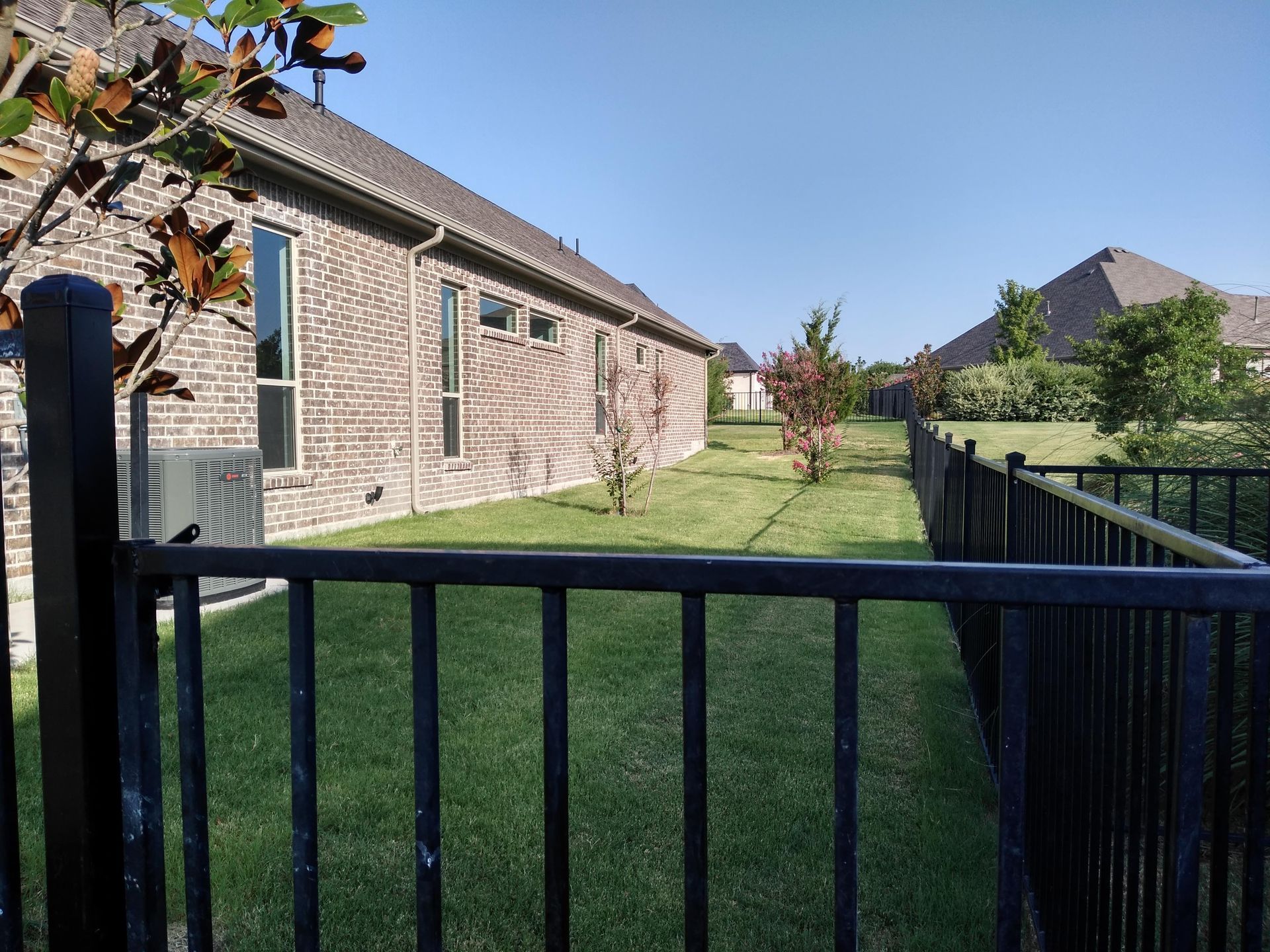 A black fence surrounds a lush green yard in front of a brick house.