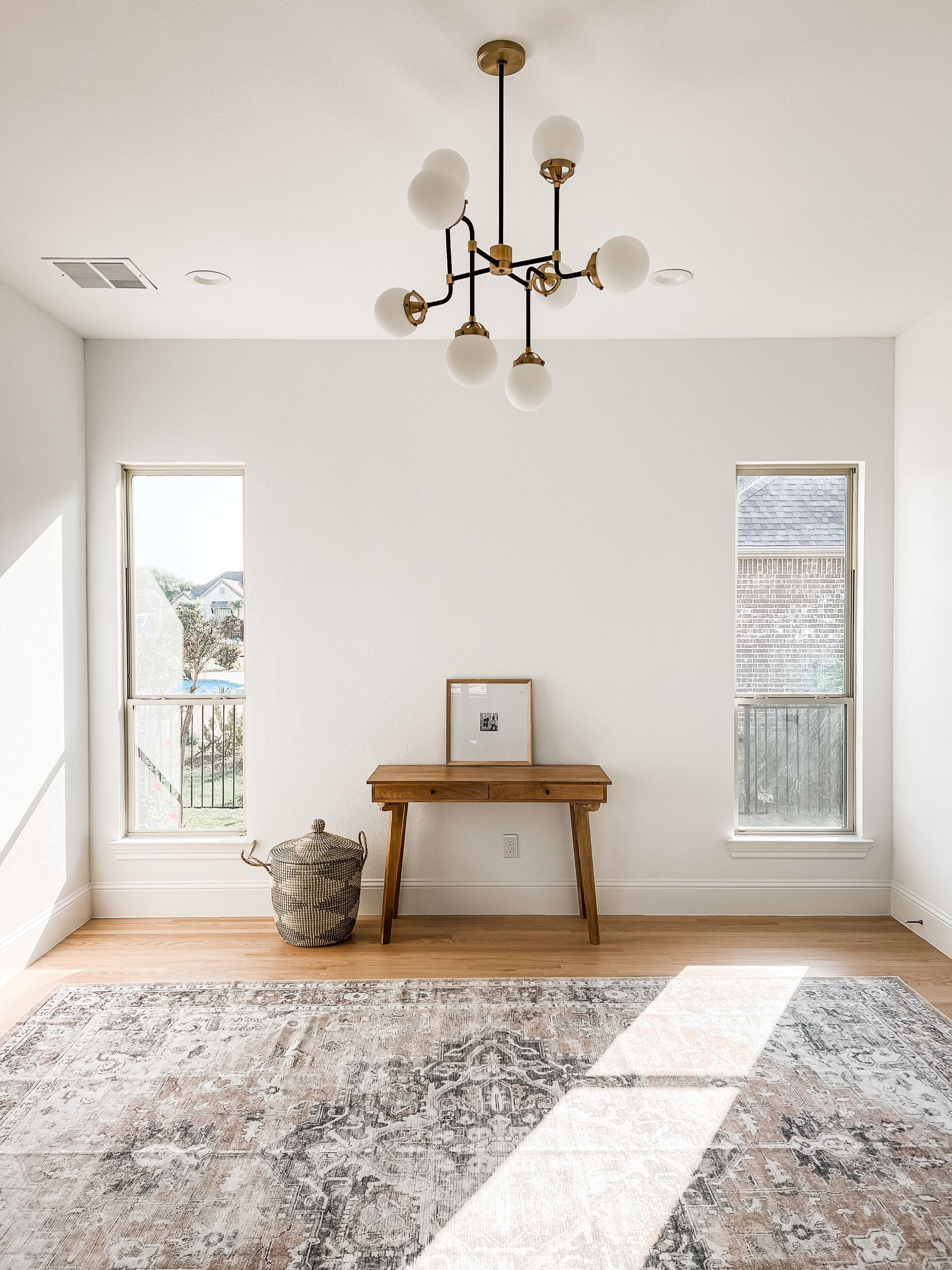 A living room with a rug , a table , a chandelier and two windows.