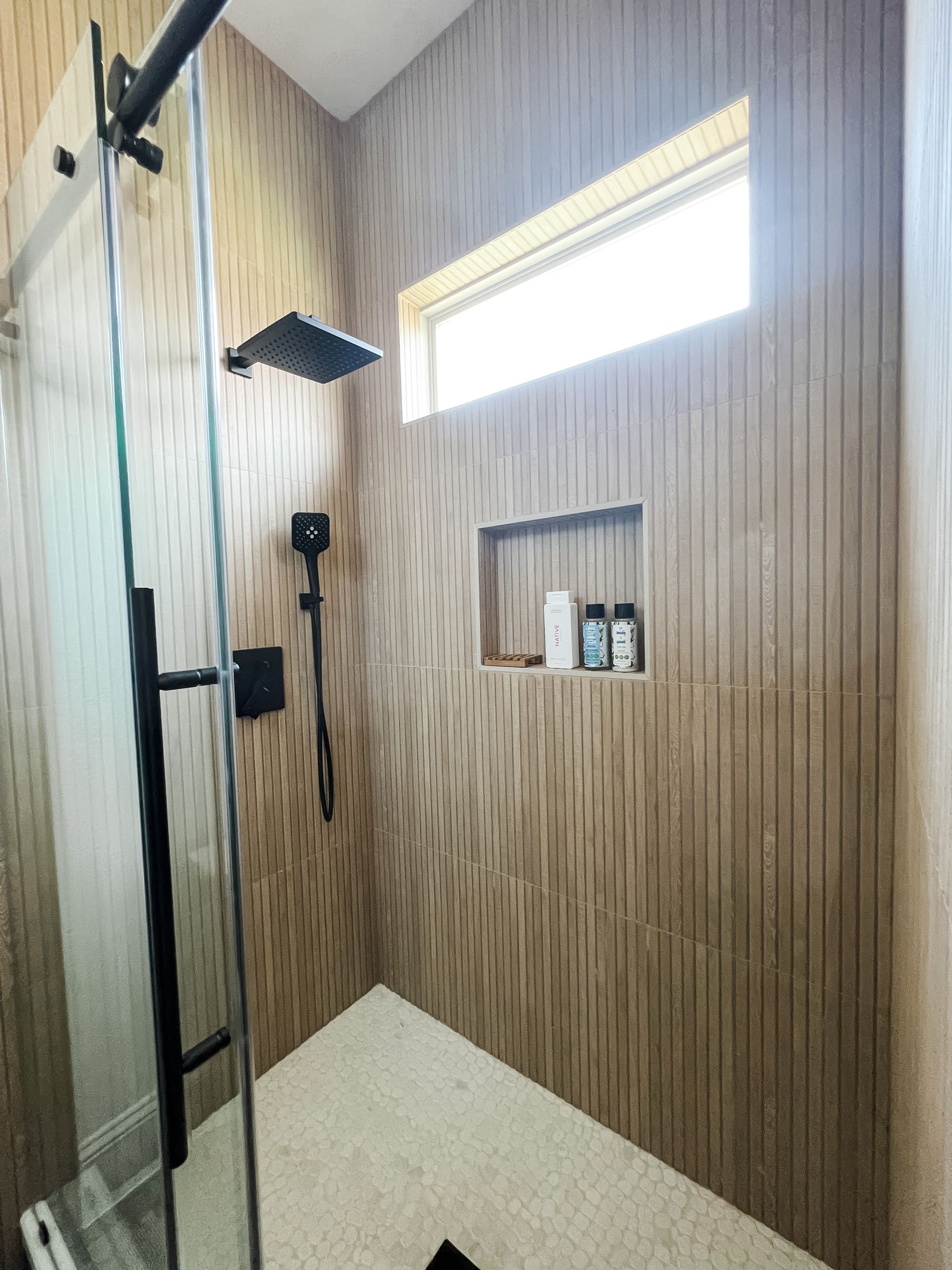Modern shower with vertical wood-look walls, black fixtures, glass door, and tiled floor. A small window and a recessed shelf are also present.