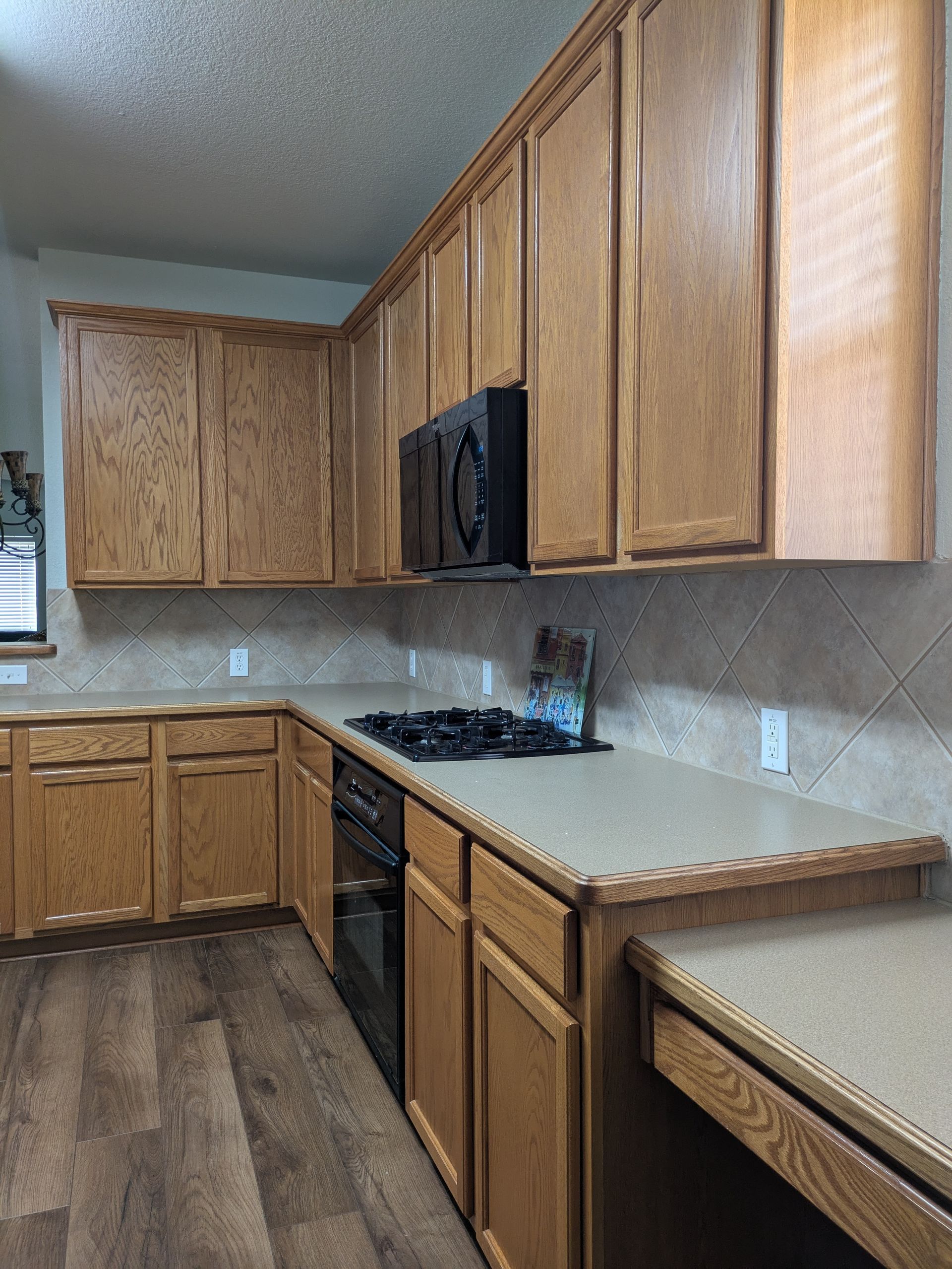 Kitchen with light wood cabinets, beige countertops, and a black microwave.