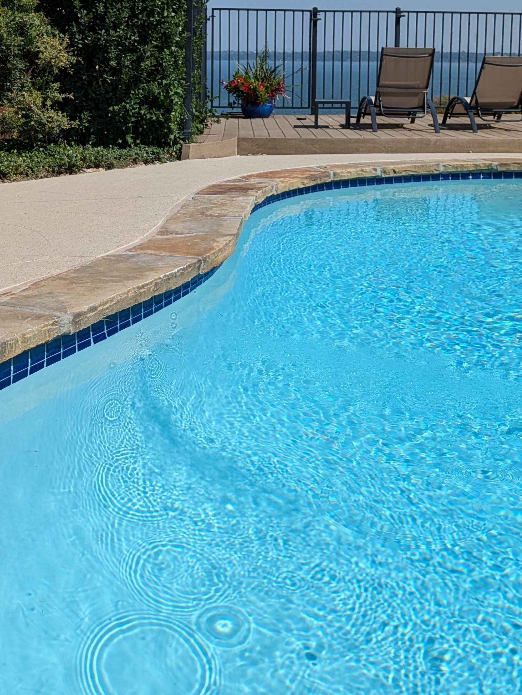 A large swimming pool surrounded by chairs and a fence with a view of the ocean.