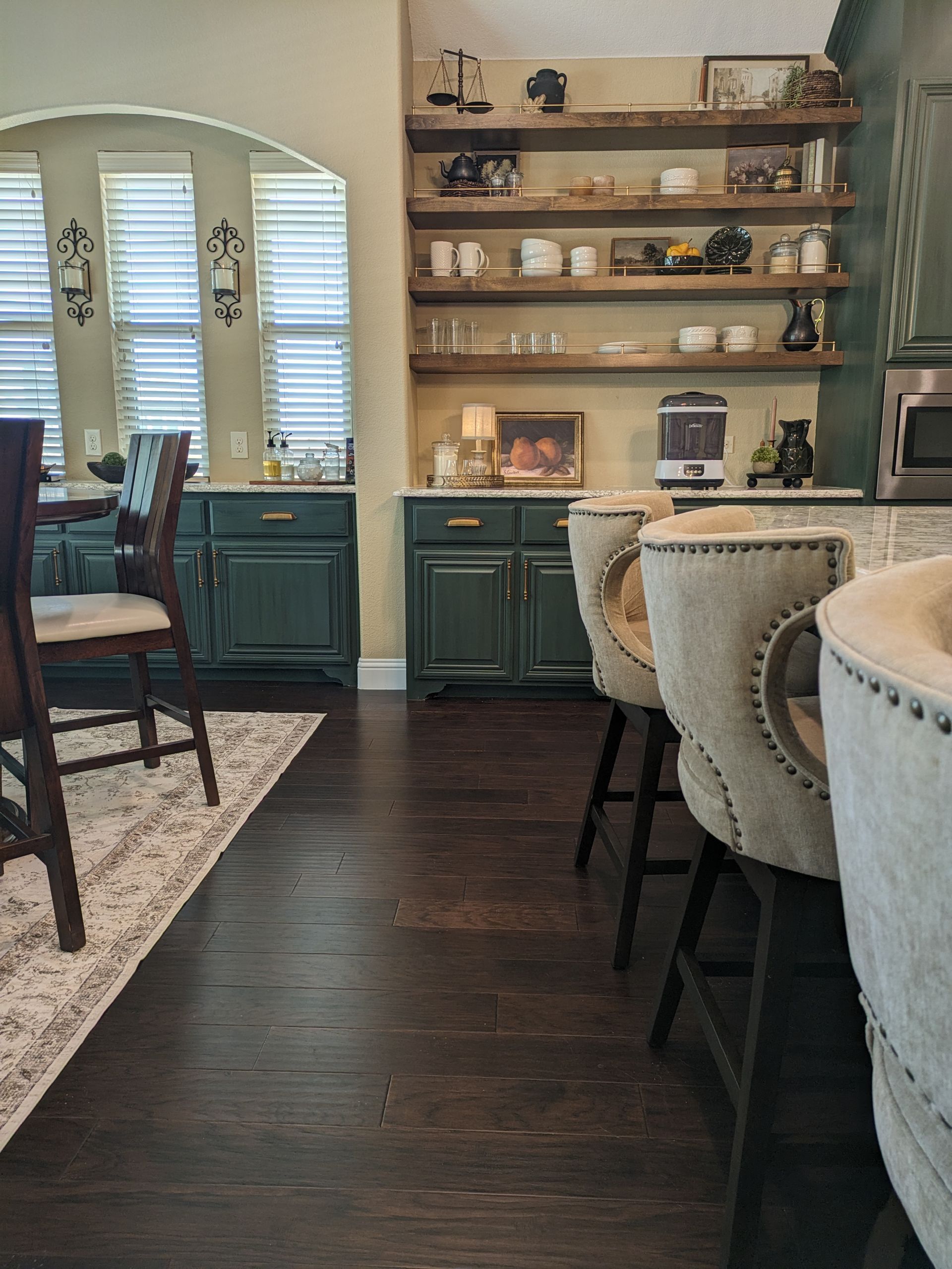 Dark wood-floored kitchen with dark green cabinets, open shelves, and a breakfast bar with high chairs.