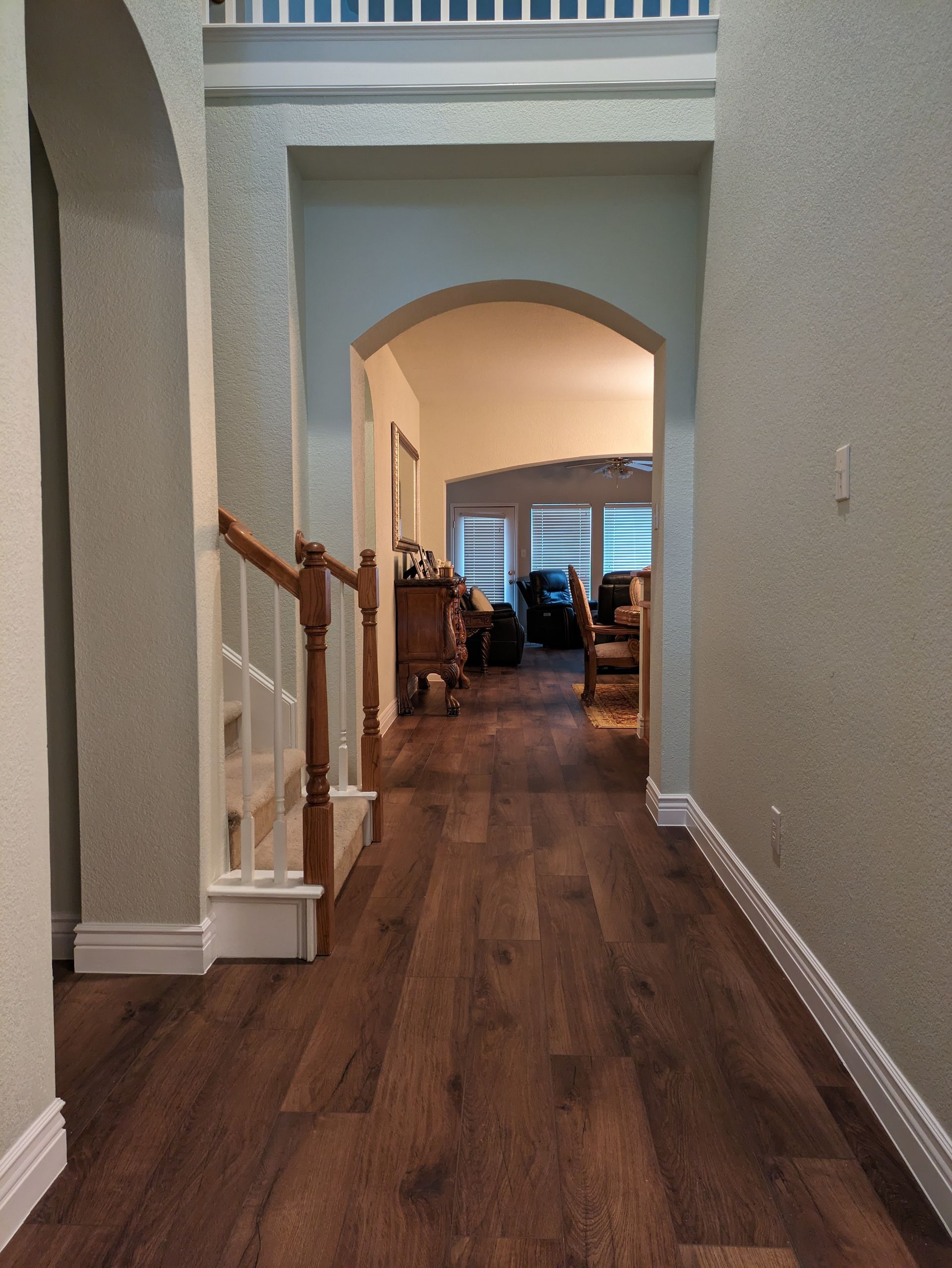 Hallway with wood floors leading to a living room; staircase on left, arched doorways, light walls.