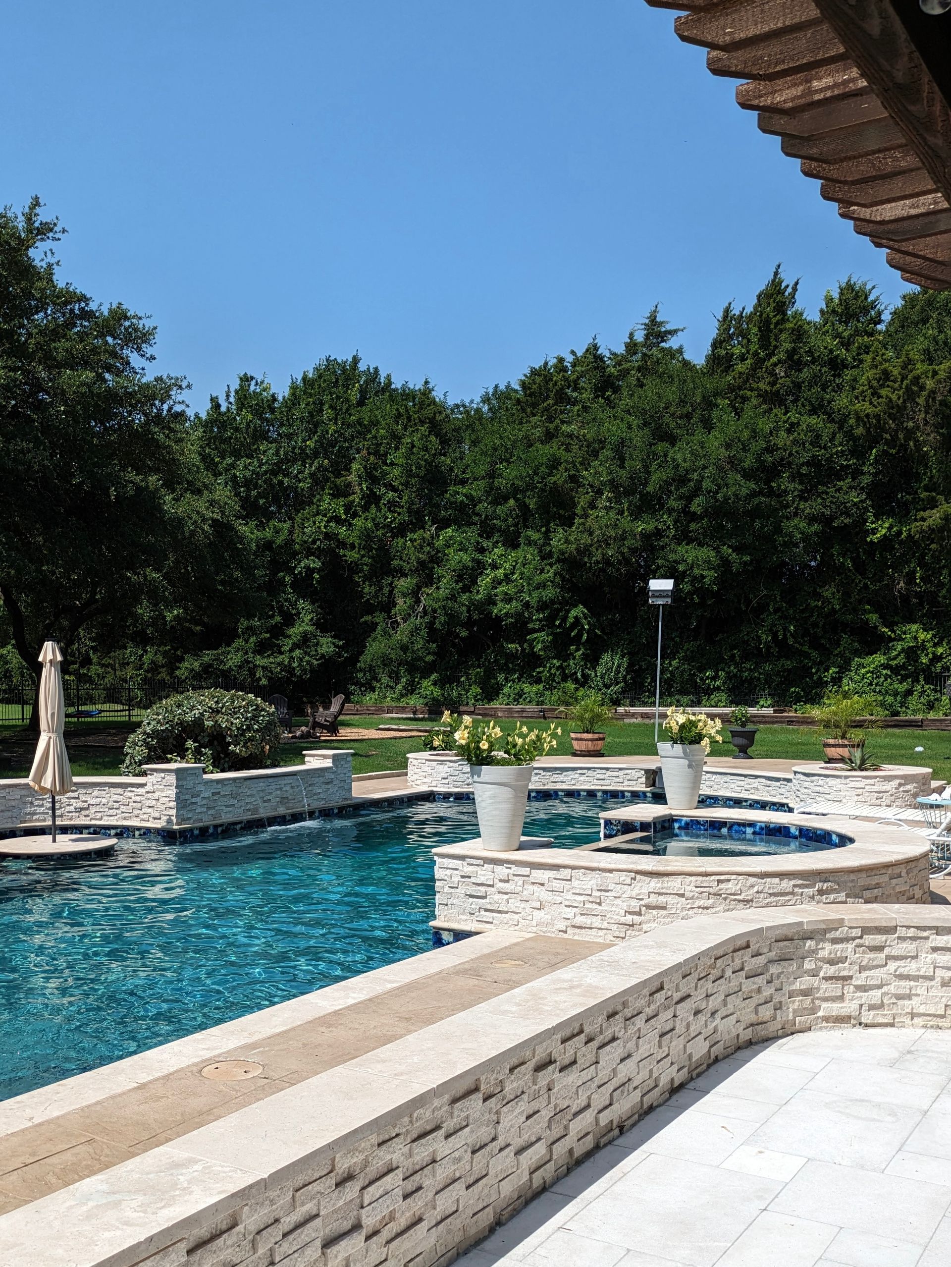 Swimming pool with clear blue water and white stone features, surrounded by trees under a clear sky.