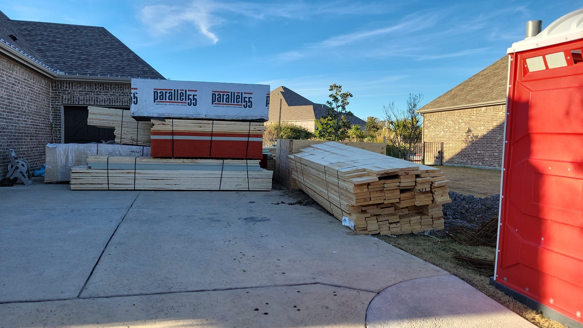 A stack of wood is sitting in a driveway next to a red portable toilet.