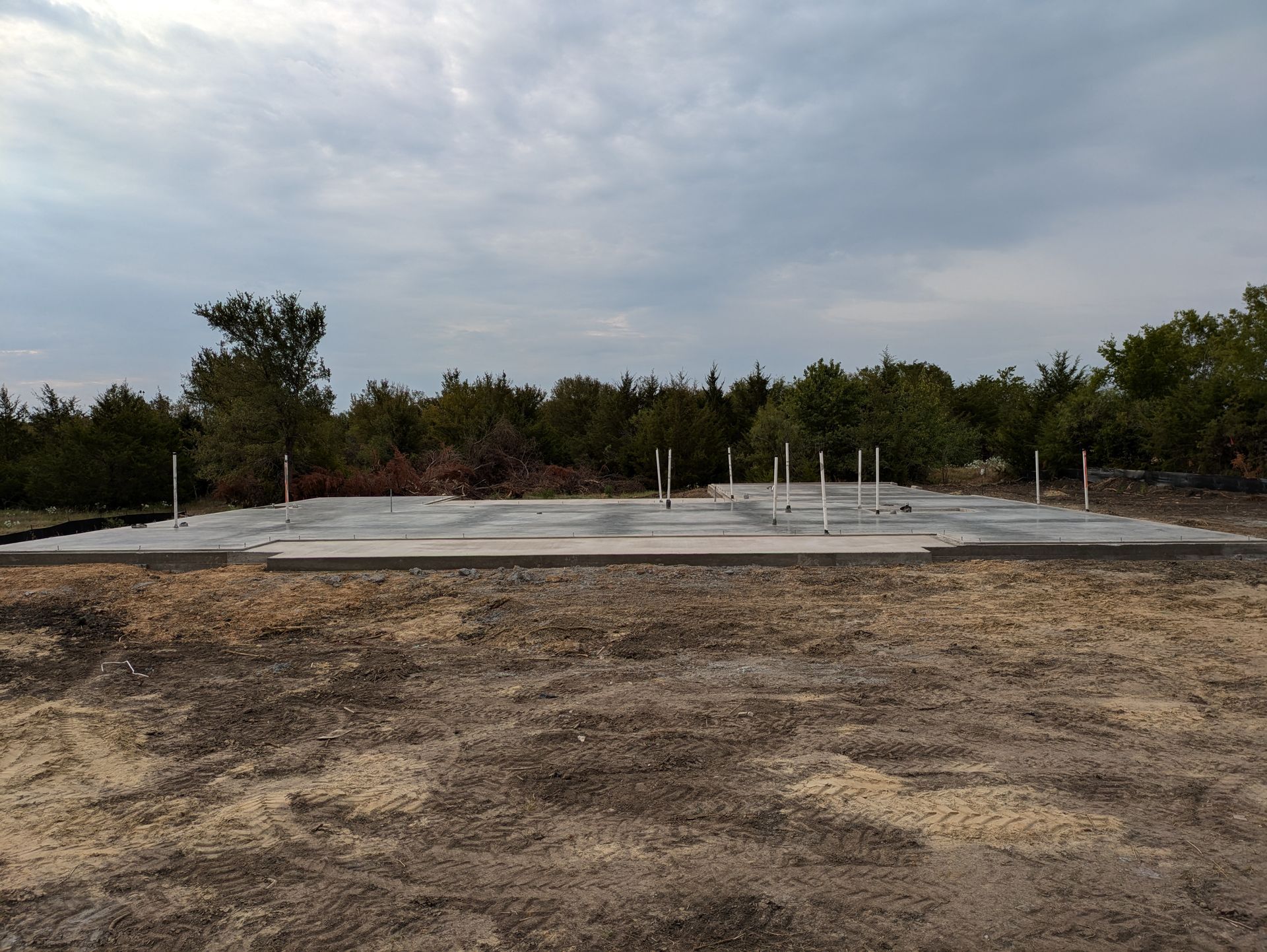 Concrete foundation with PVC pipes, in a clearing before a tree line under a cloudy sky.