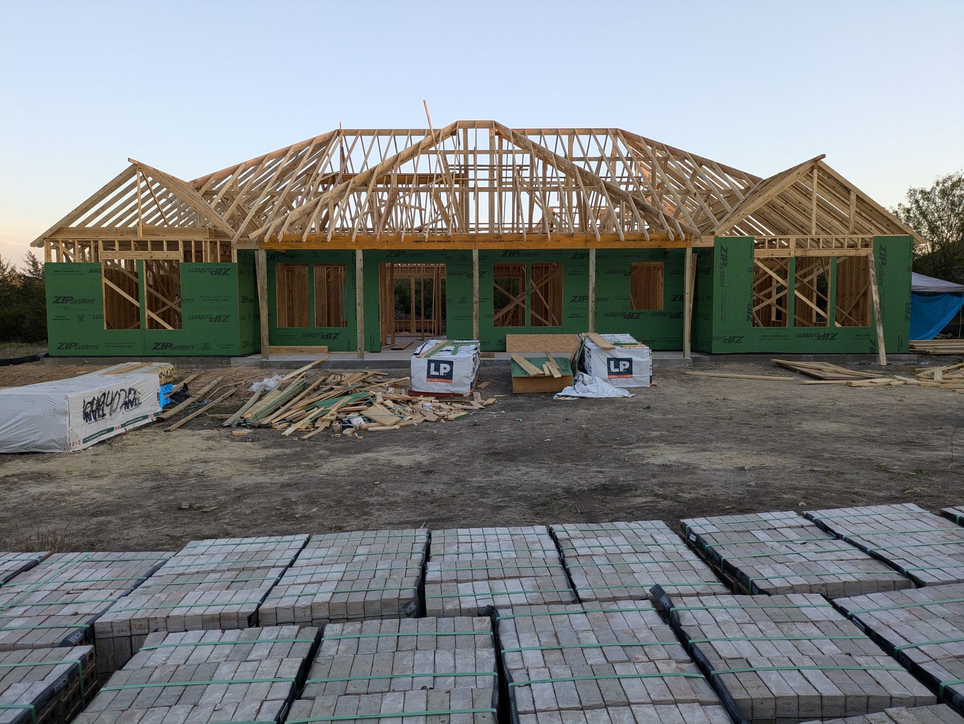 House under construction; wood framing and green sheathing visible. Bricks stacked in foreground.