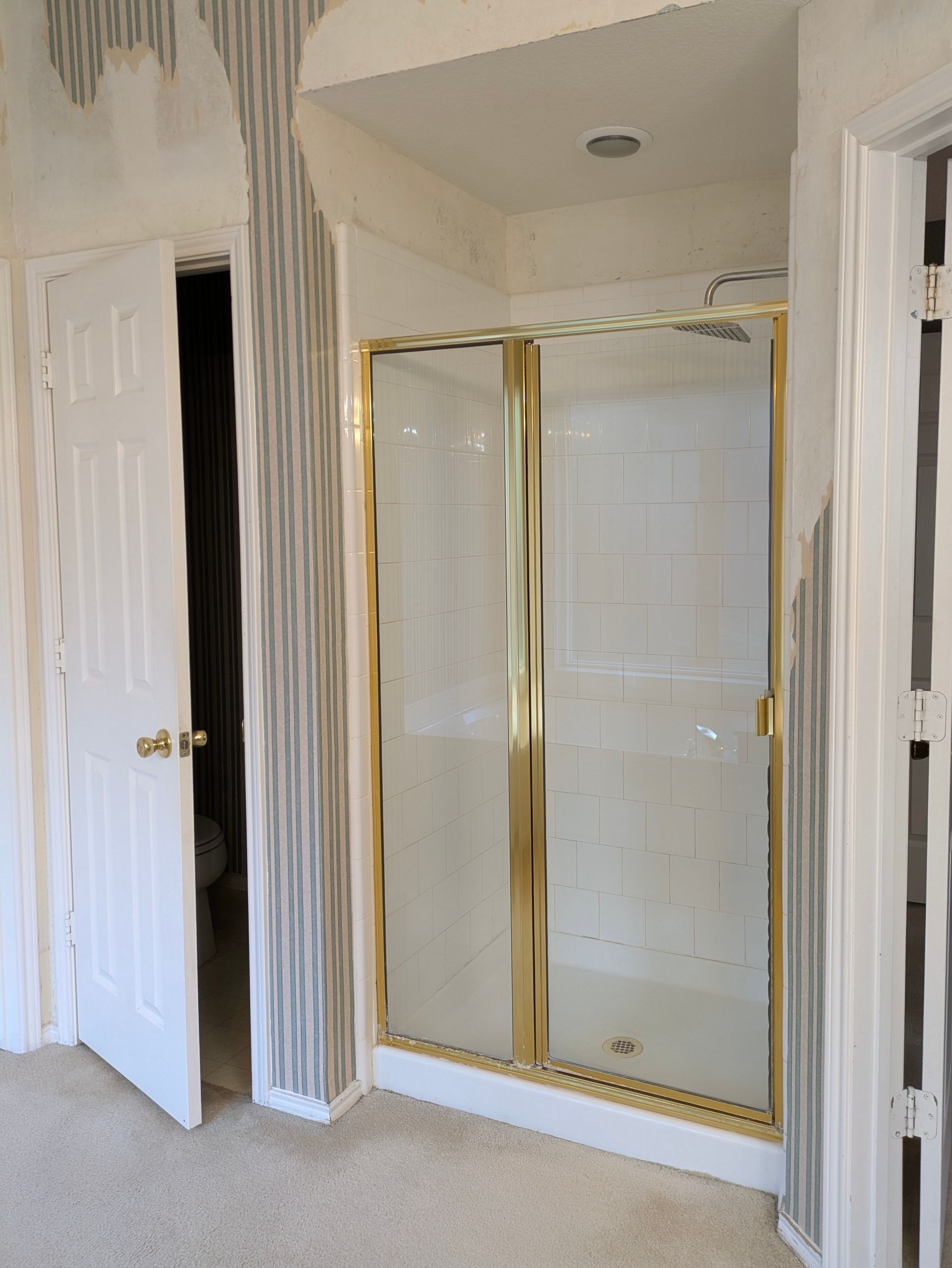 A bathroom with a shower and partially open door. Gold framed shower doors. Beige carpet.