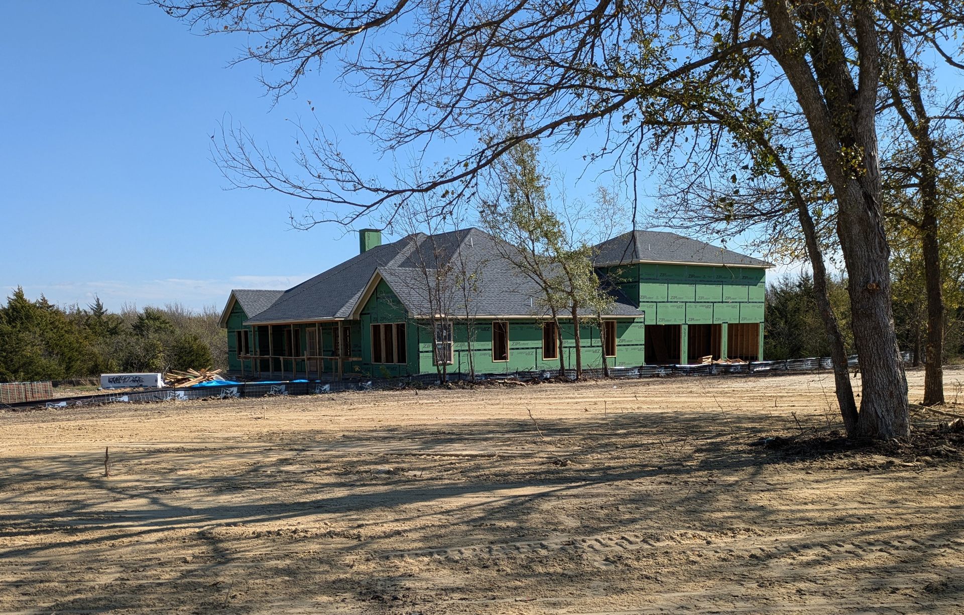 House under construction with green wrap, a gray roof, and surrounding trees in a sunny field.