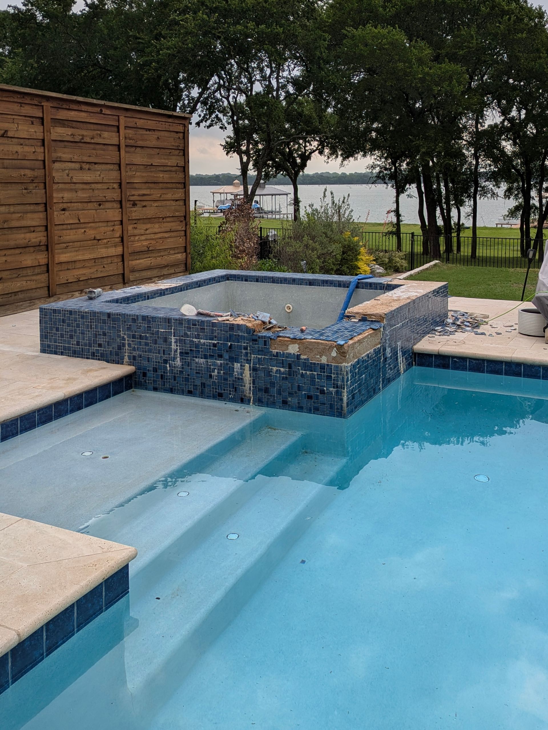 Pool with blue tile, steps, and spa. A wooden fence and lake are visible in the background.