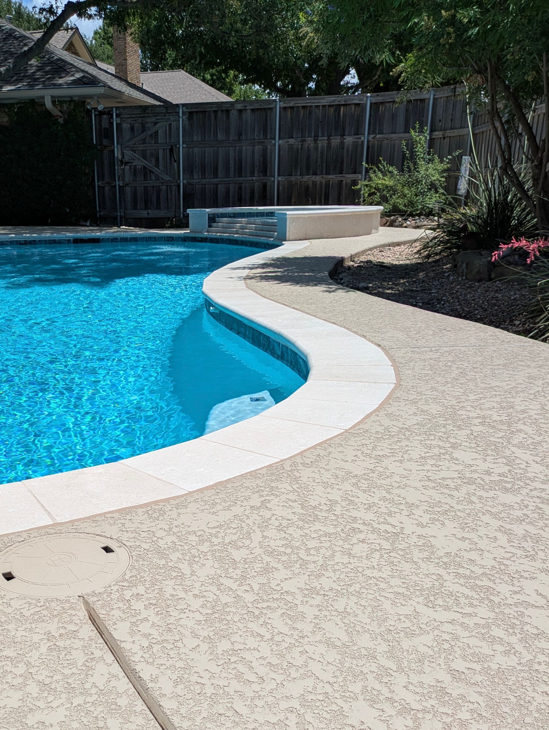 Swimming pool with blue water, surrounded by a light-colored concrete deck. A low wall is near the pool.