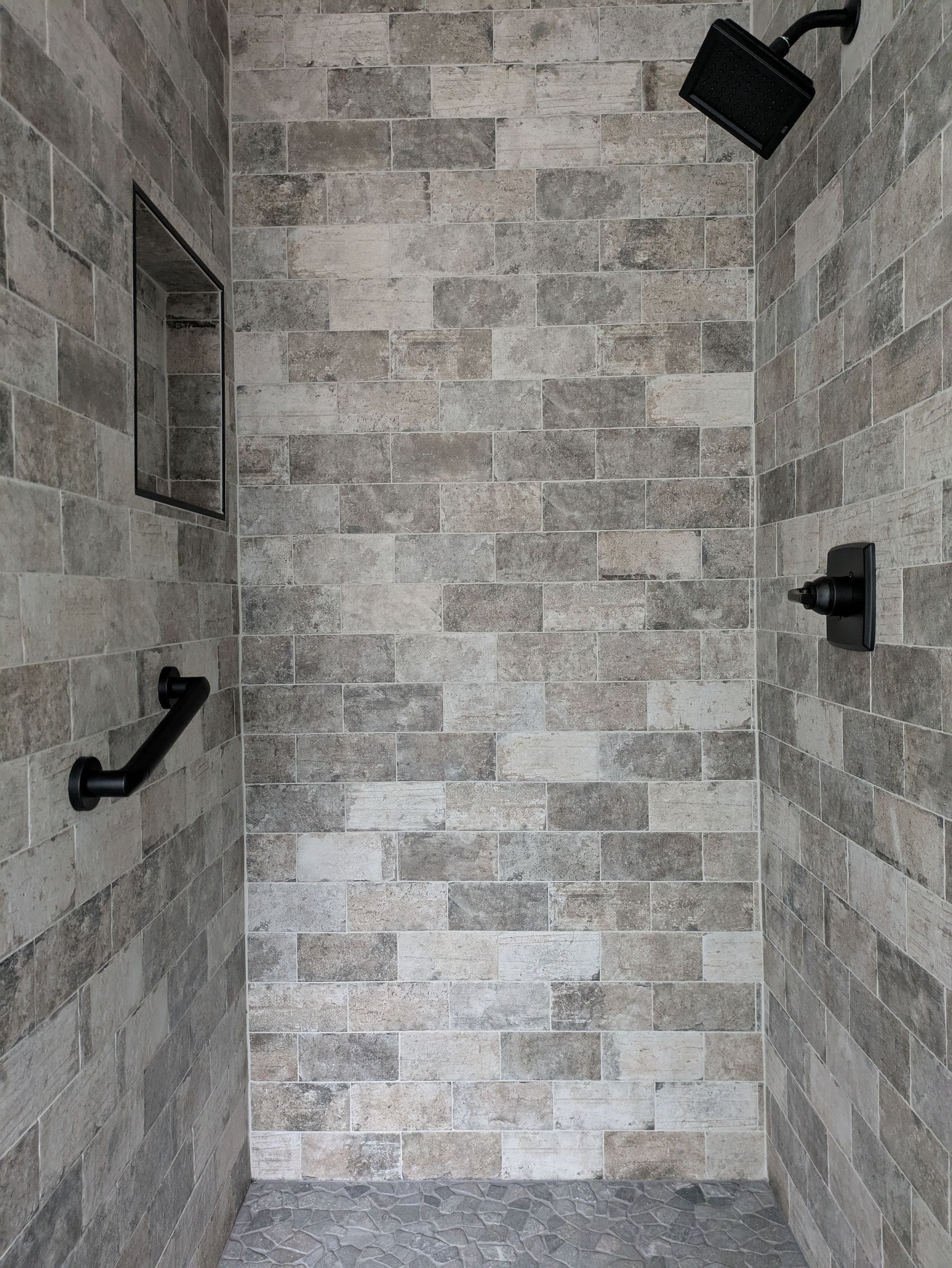 Shower stall with gray stone tile, shower head, faucet, and grab bar.