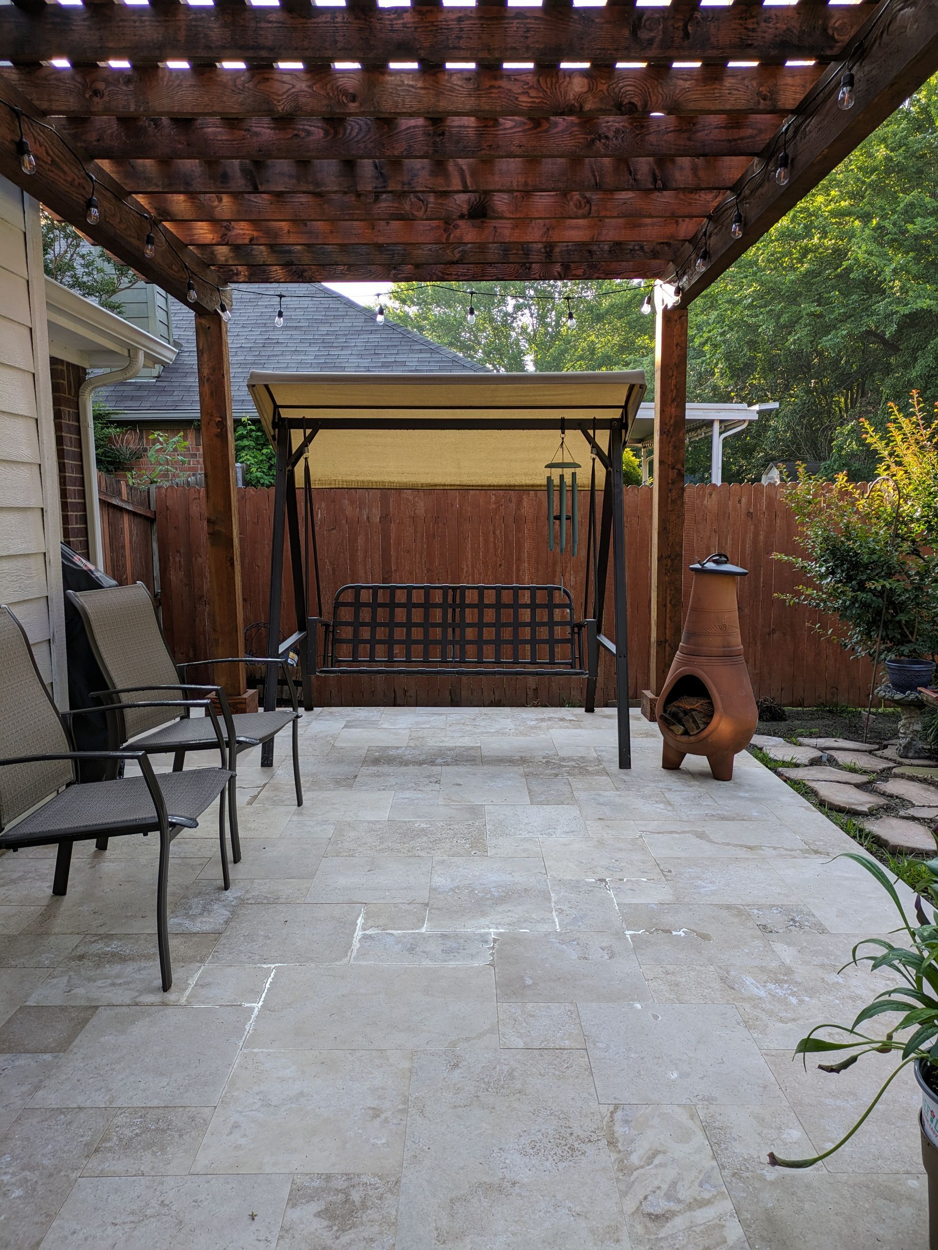 Patio with pergola, swing, chairs, and fire pit. Brown wood, tan roof, stone patio.
