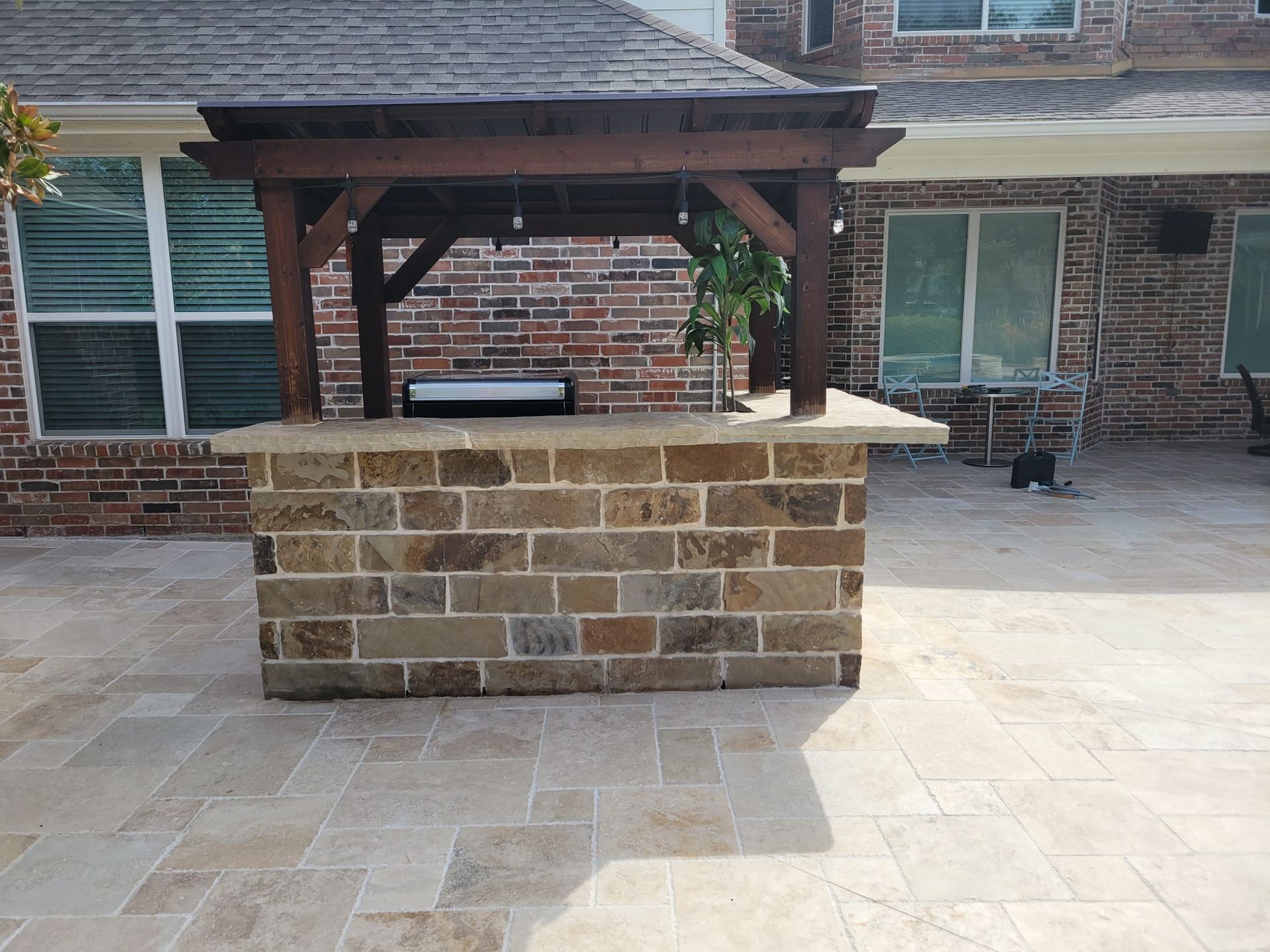 A brick island with a grill under a wooden pergola on a patio.